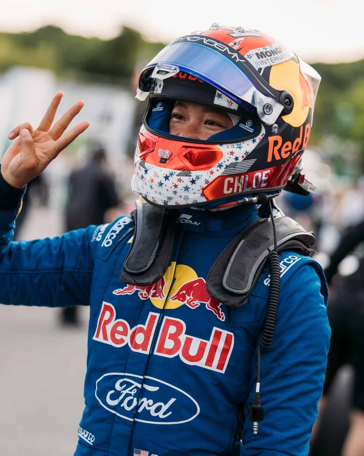 A race car driver wearing a Red Bull and Ford racing suit and helmet smiles and holds up a peace sign with their fingers.
