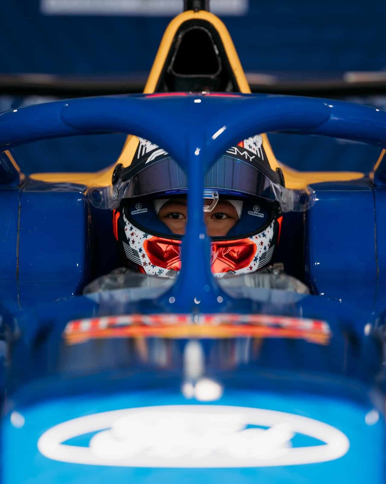 A race car driver, wearing a helmet and red reflective visor, sits in a blue open-wheel race car with a visible Ford logo on the nose. The driver is focused and ready for action.