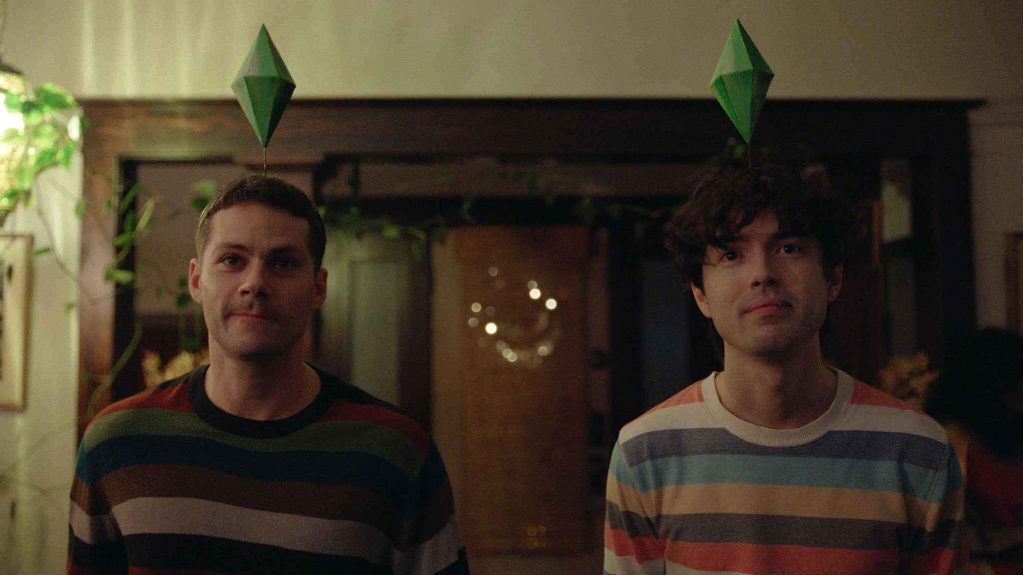 Two people wearing striped sweaters stand indoors, each with a green Sims-style plumbob headpiece. The background shows a doorway with plants and warm lighting, creating a casual, playful atmosphere.