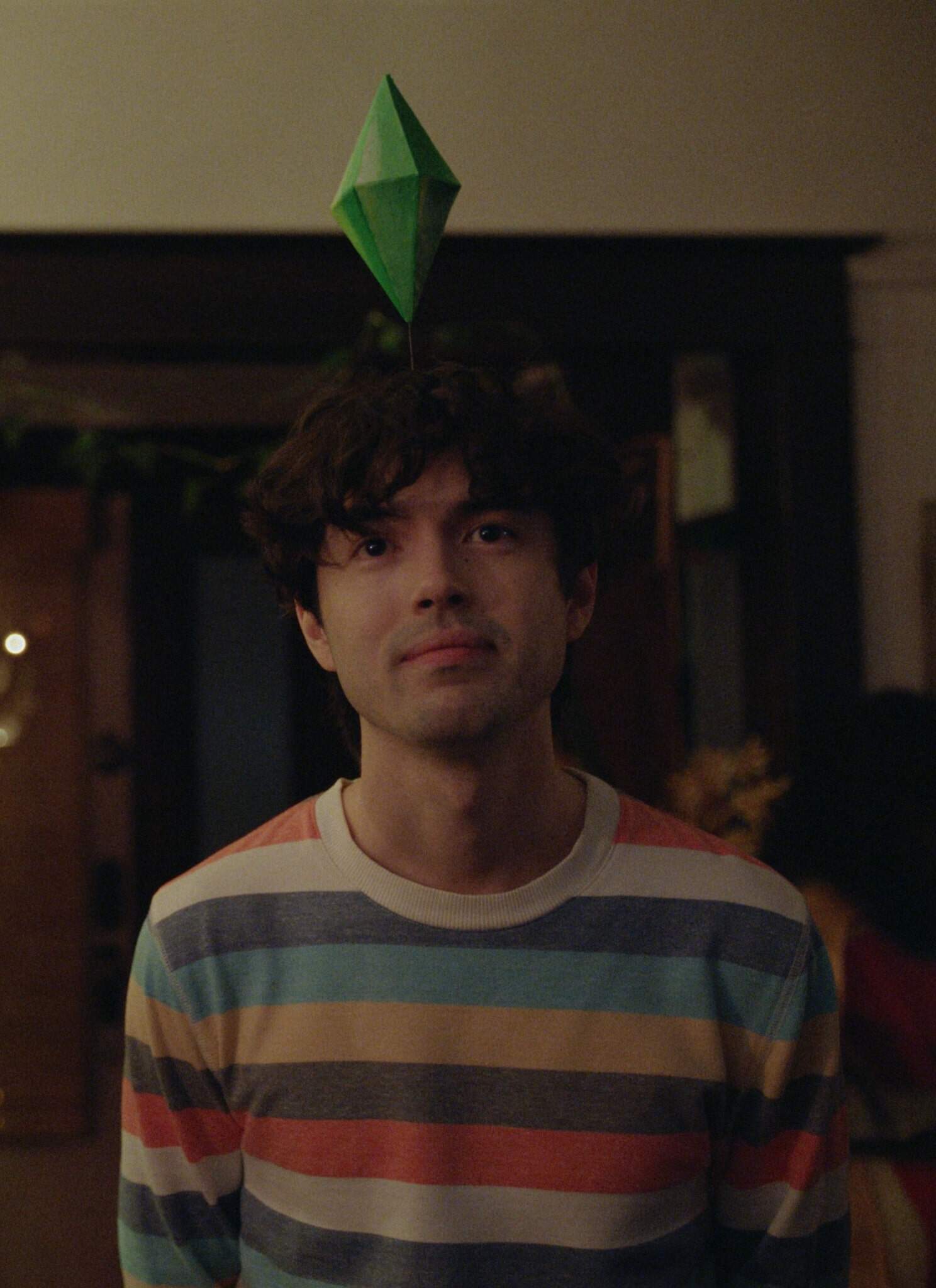 Two people wearing striped sweaters stand indoors, each with a green Sims-style plumbob headpiece. The background shows a doorway with plants and warm lighting, creating a casual, playful atmosphere.