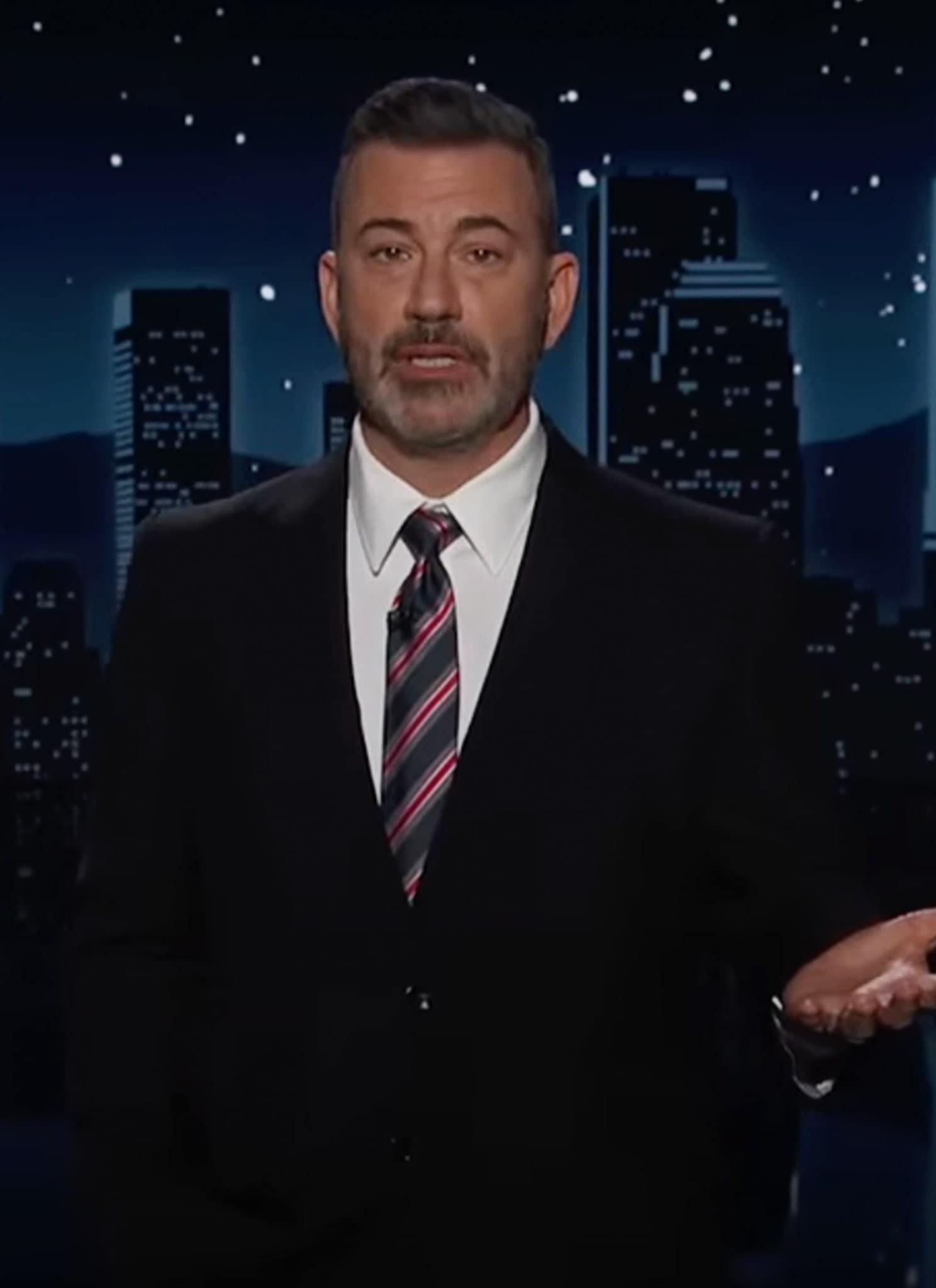 A man in a suit and striped tie stands in front of a nighttime city skyline backdrop, speaking with one hand raised, as if hosting a late-night television show.