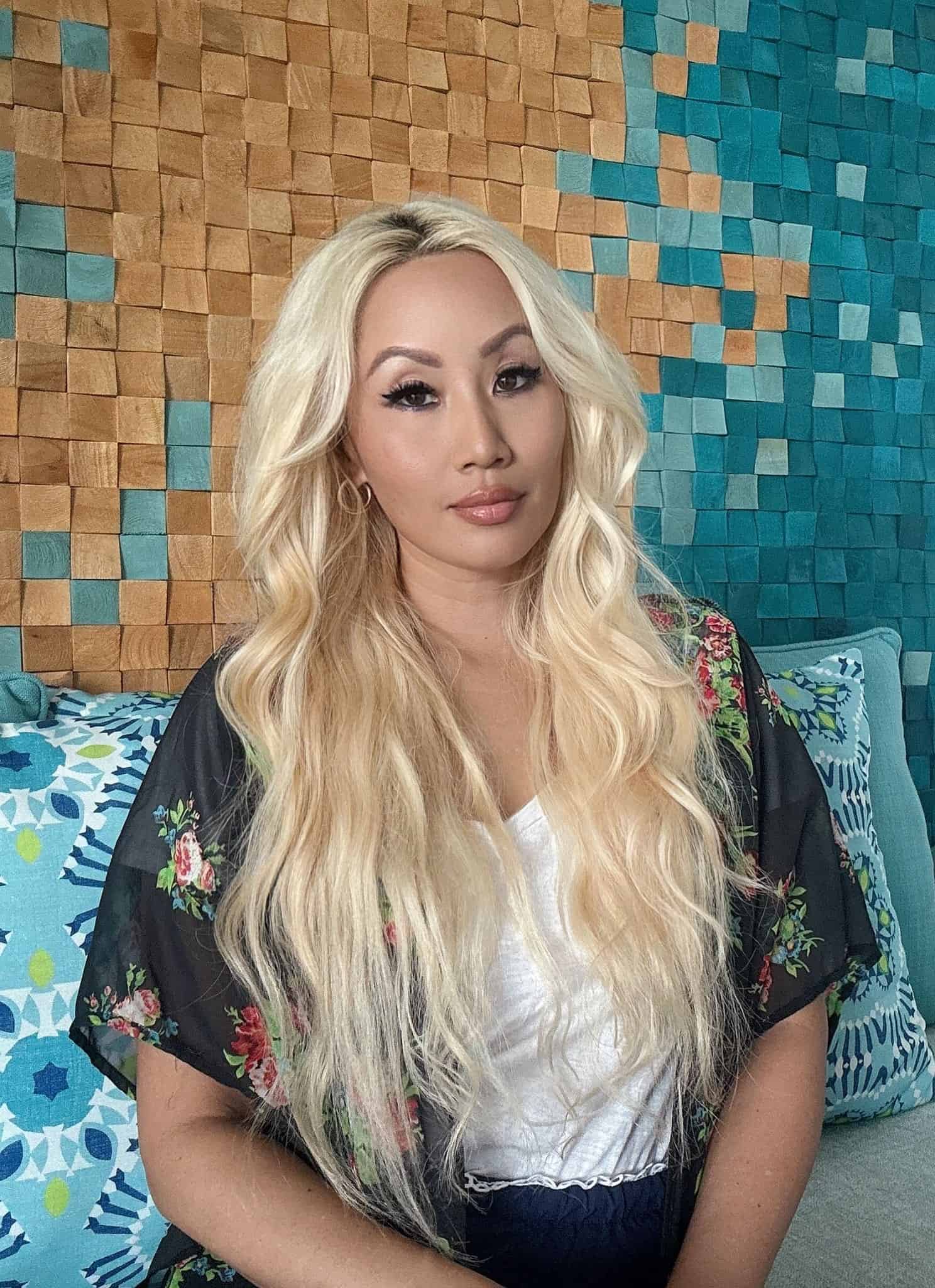 A woman with long blonde hair sits on a couch with teal and blue cushions. Next to her is a book cover titled Fetishized: A Reckoning with Yellow Fever, Feminism, and Beauty by Kaila Yu, featuring a black and white photo of a woman.