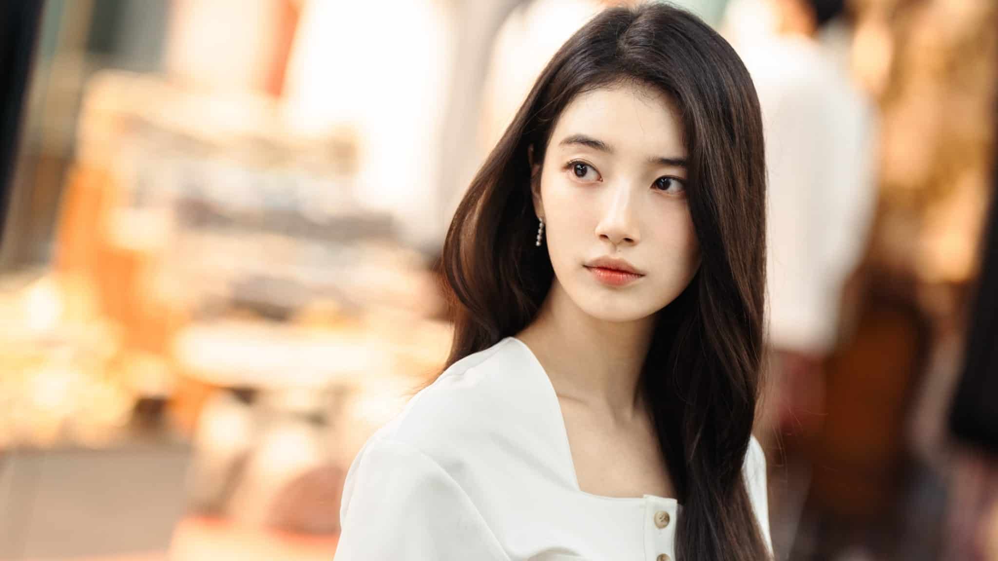 A young woman with long dark hair wearing a white blouse looks to the side in a softly lit indoor setting. The background is blurred, focusing attention on her thoughtful expression.