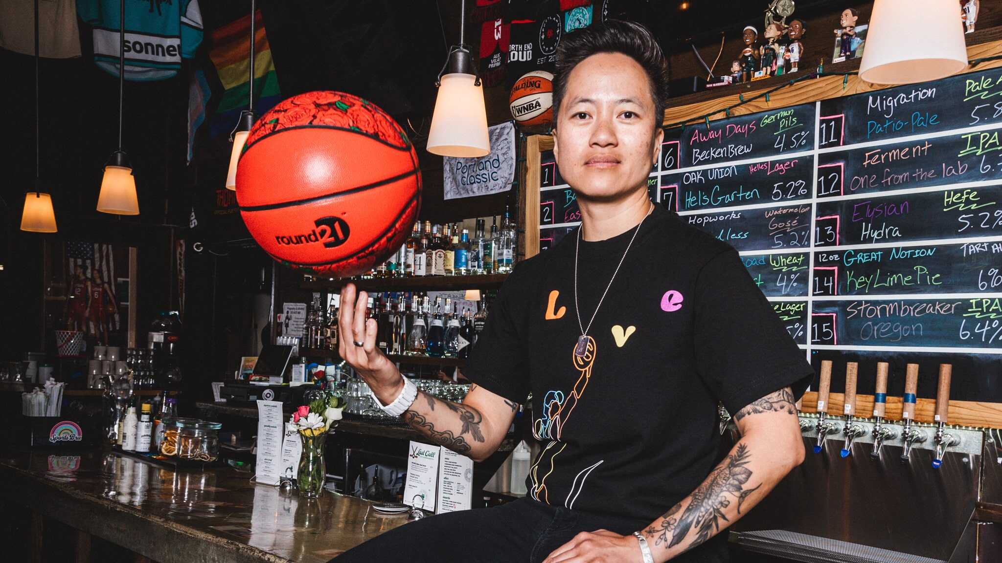 A person sits on a bar counter, spinning a red basketball on one finger. They wear a black t-shirt with colorful designs and bracelets. Behind them is a chalkboard beer menu and a variety of bar decorations.