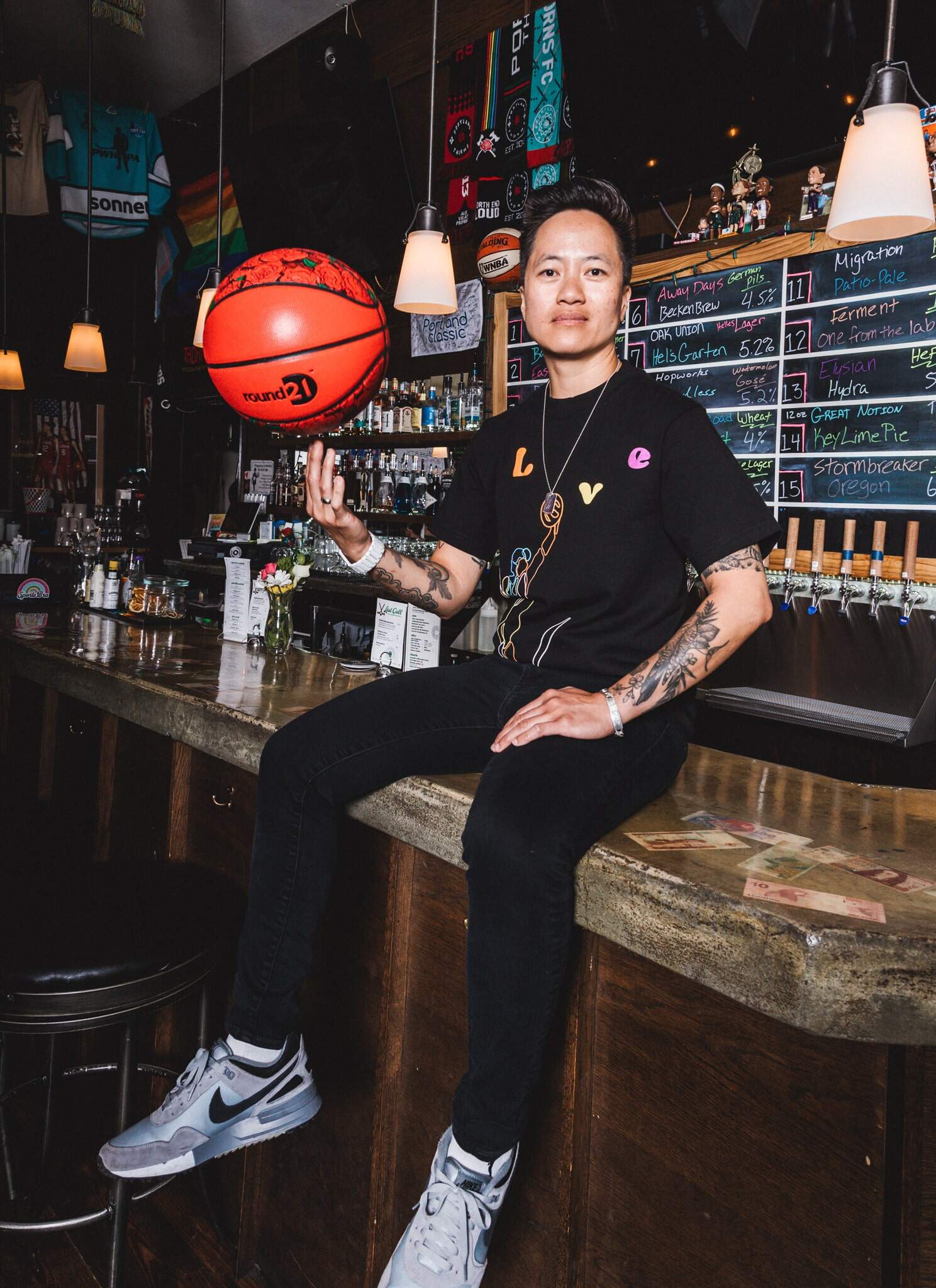 A person sits on a bar counter, spinning a red basketball on one finger. They wear a black t-shirt with colorful designs and bracelets. Behind them is a chalkboard beer menu and a variety of bar decorations.