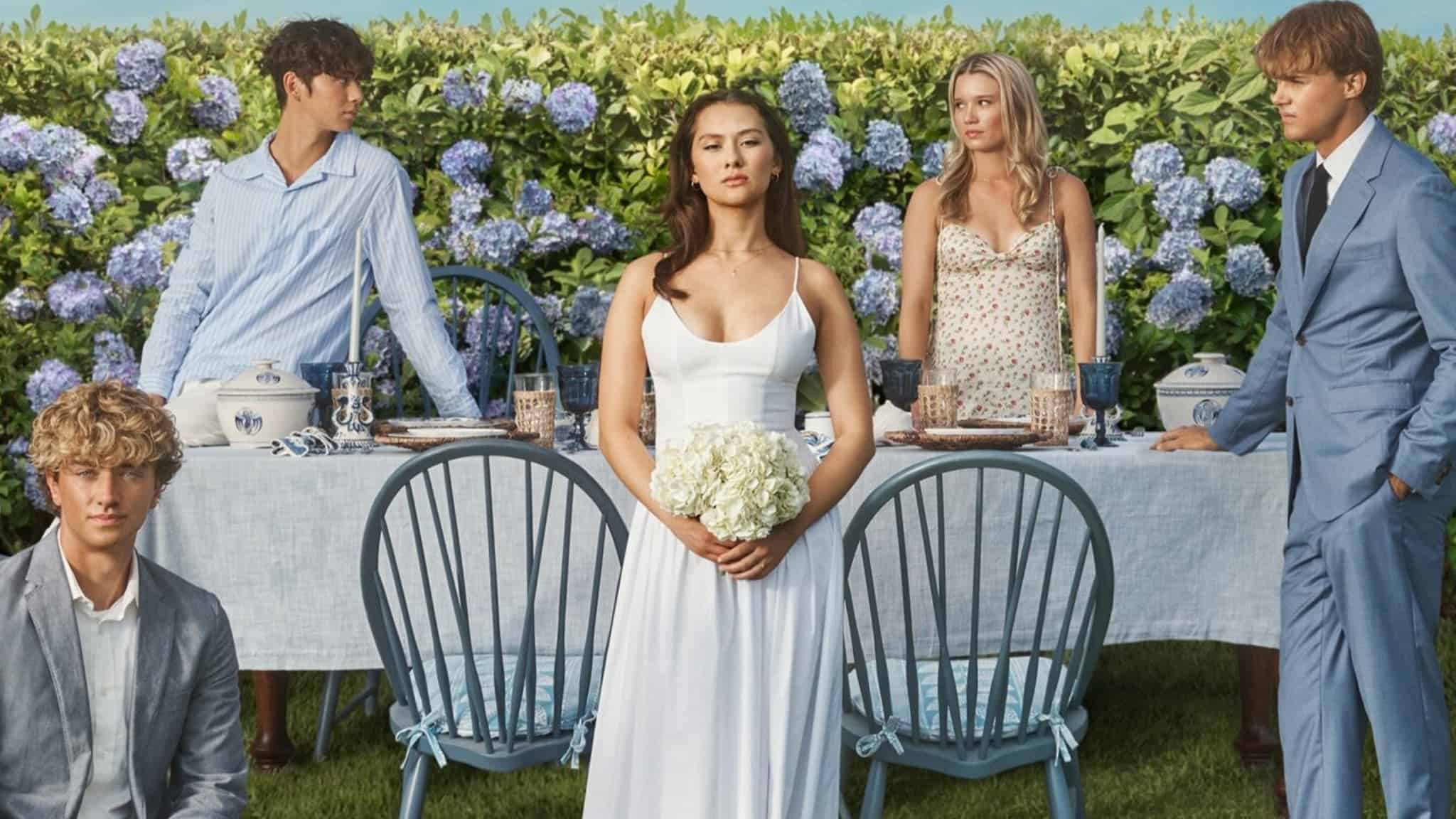 A young woman in a white dress stands holding flowers in front of a garden table, surrounded by four young adults dressed in pastel summer outfits, with a lush hedge of blue and purple hydrangeas behind them.