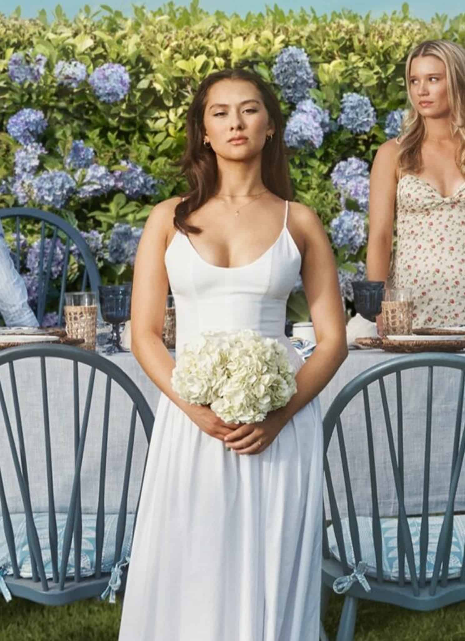 A young woman in a white dress stands holding flowers in front of a garden table, surrounded by four young adults dressed in pastel summer outfits, with a lush hedge of blue and purple hydrangeas behind them.