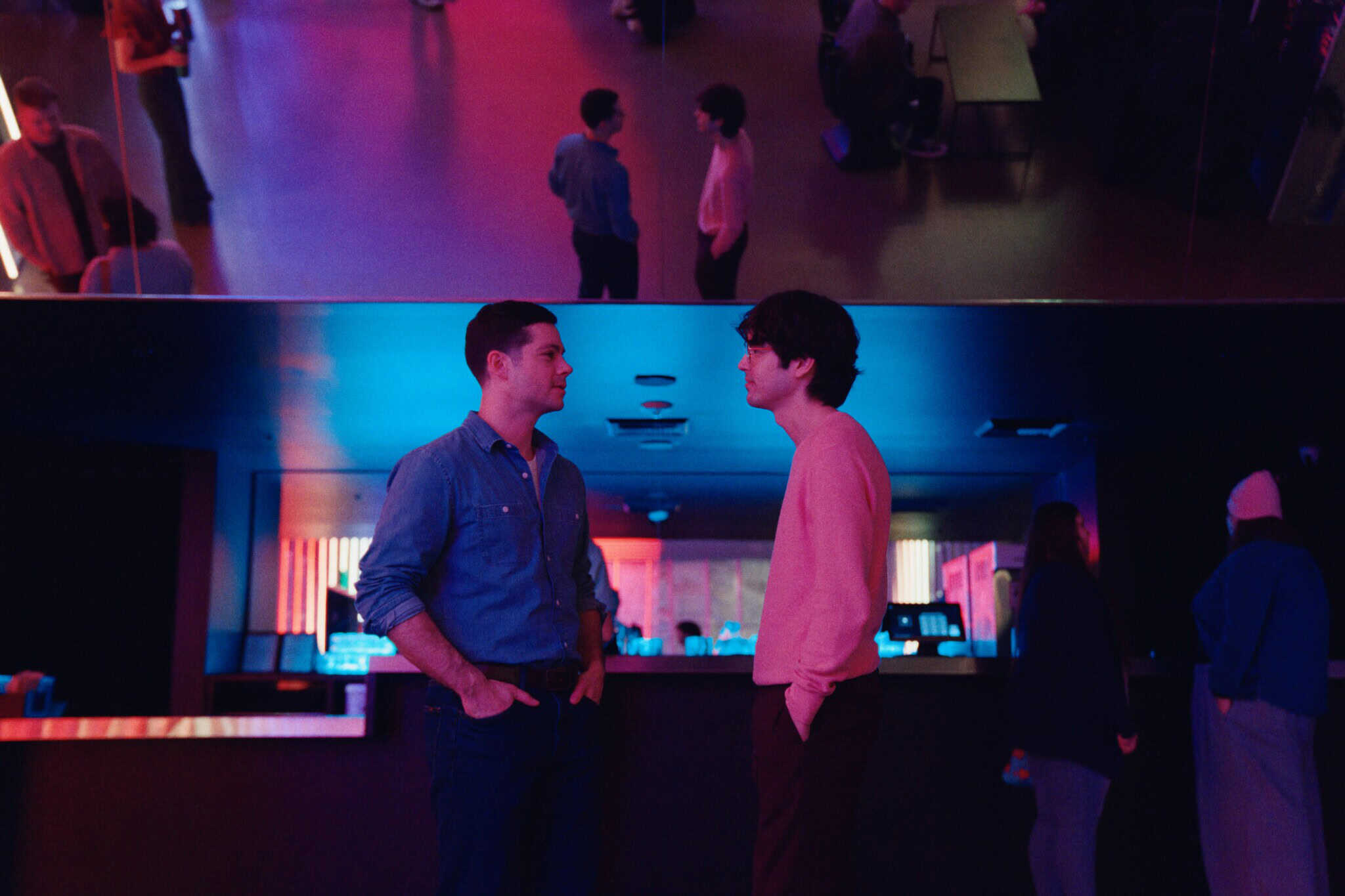 Two men stand facing each other and talking in a dimly lit room with neon pink and blue lighting. Their reflections appear in a large mirror above them, along with other people in the background.