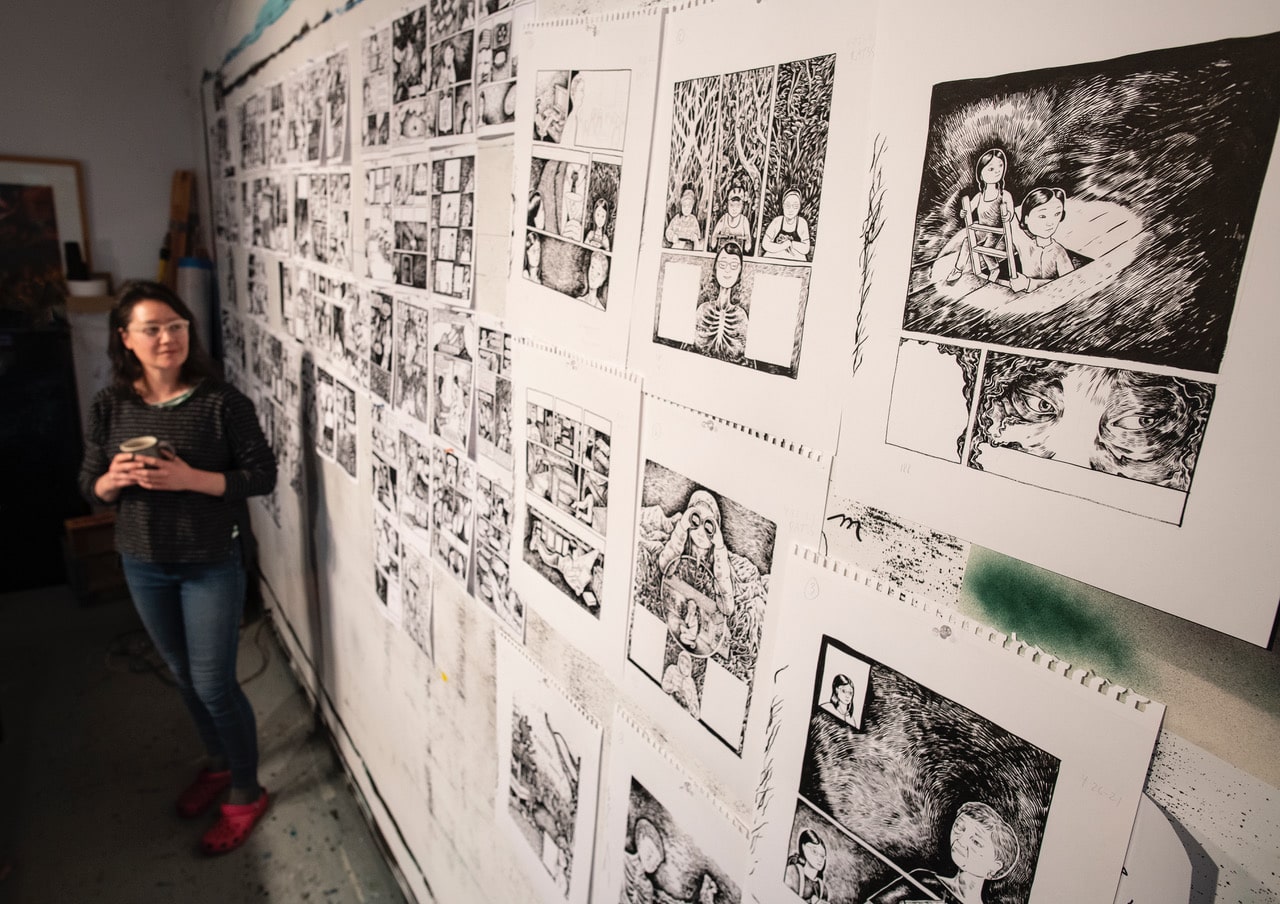 A woman stands by a wall covered with numerous black-and-white comic book pages and illustrations, holding a mug and looking at the artwork, in a creative studio setting.