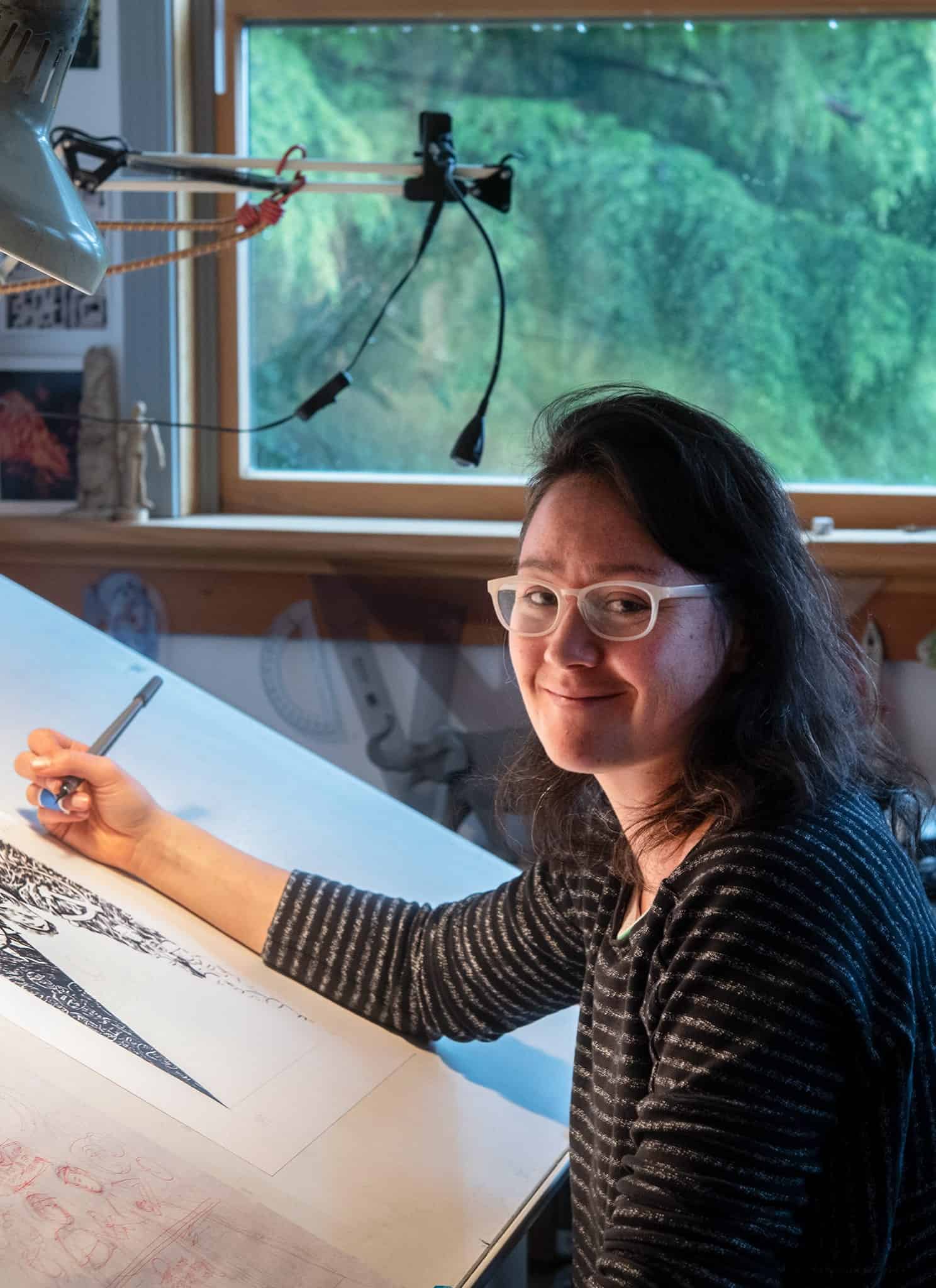 A person with glasses and dark hair smiles while drawing detailed artwork at a well-lit desk, surrounded by art supplies, near a large window with a view of greenery.