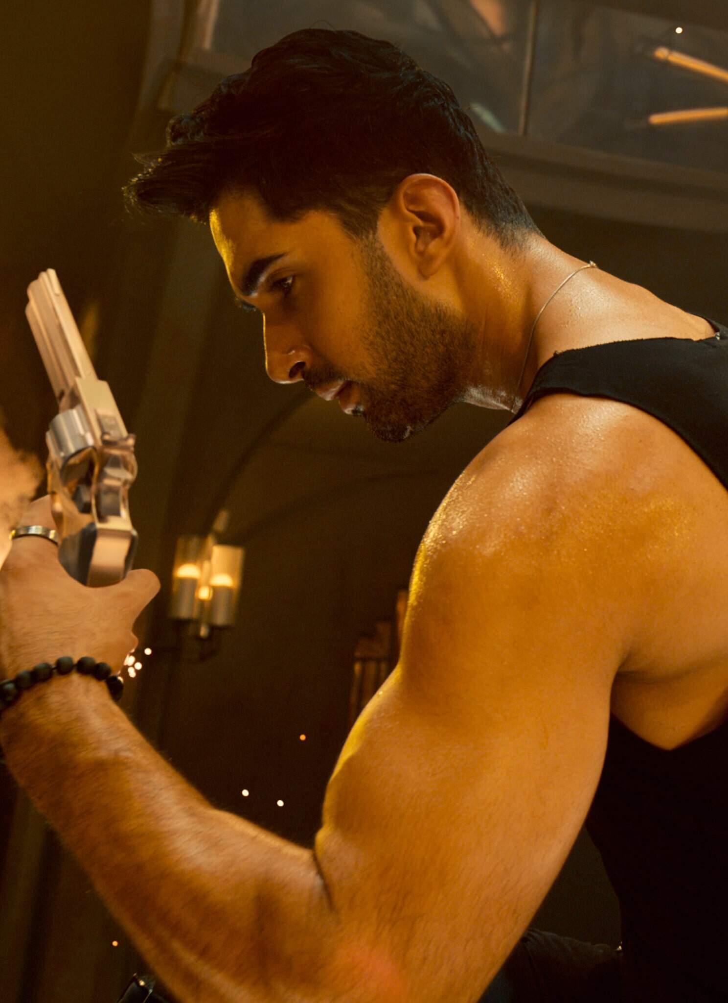 A muscular man in a black sleeveless shirt holds two silver revolvers, one pointed forward and one behind his back, with sparks flying around him in a dramatic, dimly lit setting.