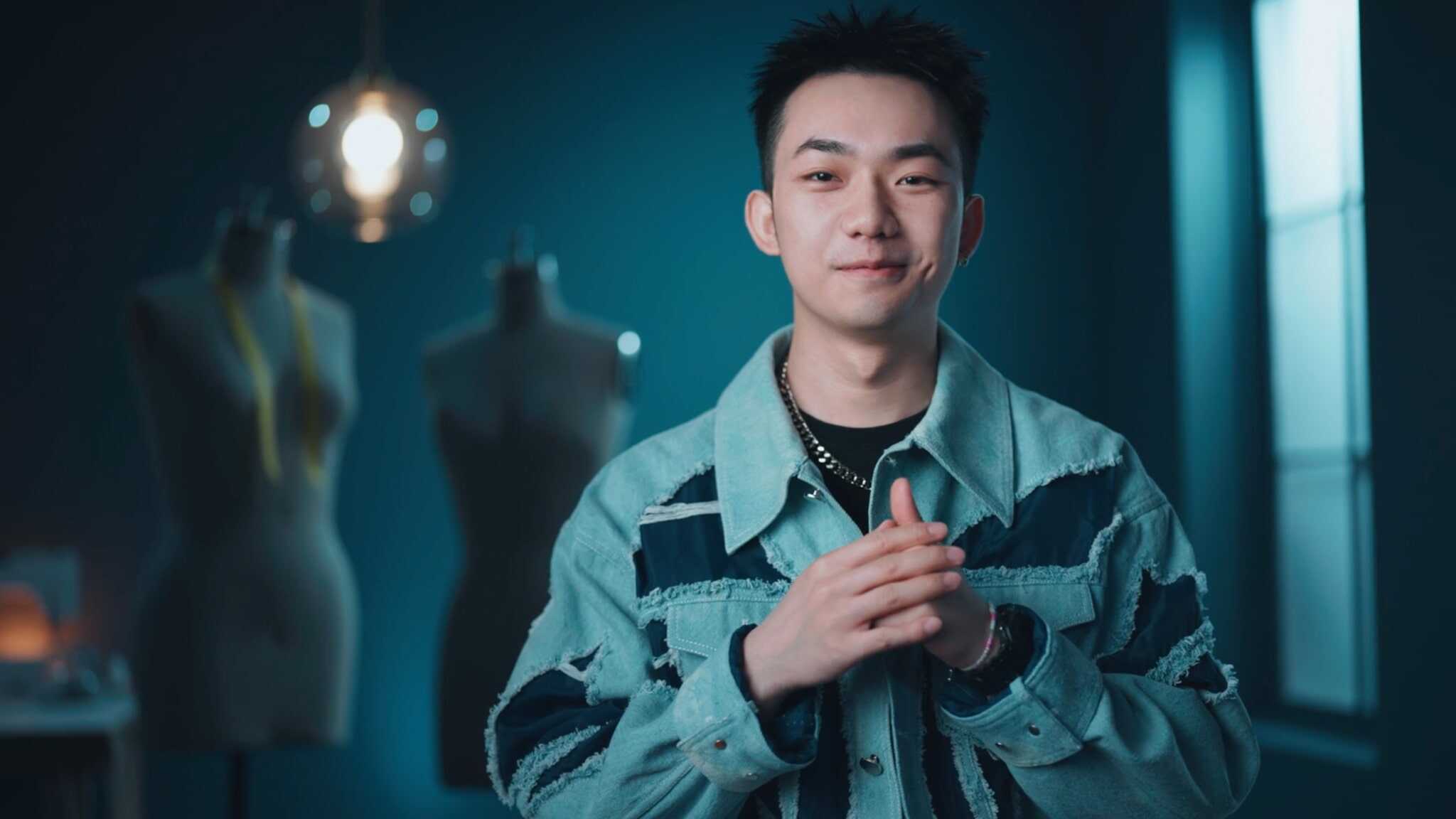 A young man wearing a blue patterned jacket stands smiling with hands clasped in front of him. Behind him are dress forms and a hanging light, in a softly lit room with blue walls.