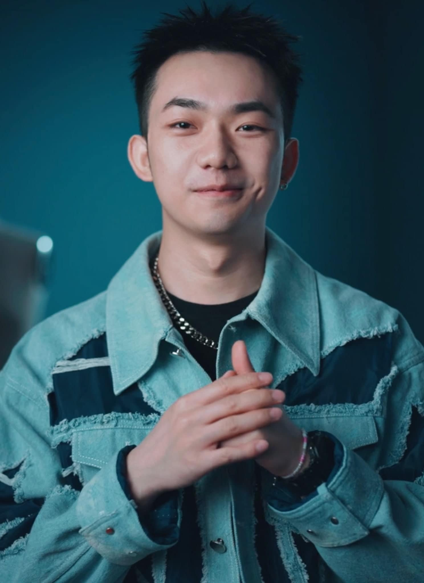A young man wearing a blue patterned jacket stands smiling with hands clasped in front of him. Behind him are dress forms and a hanging light, in a softly lit room with blue walls.