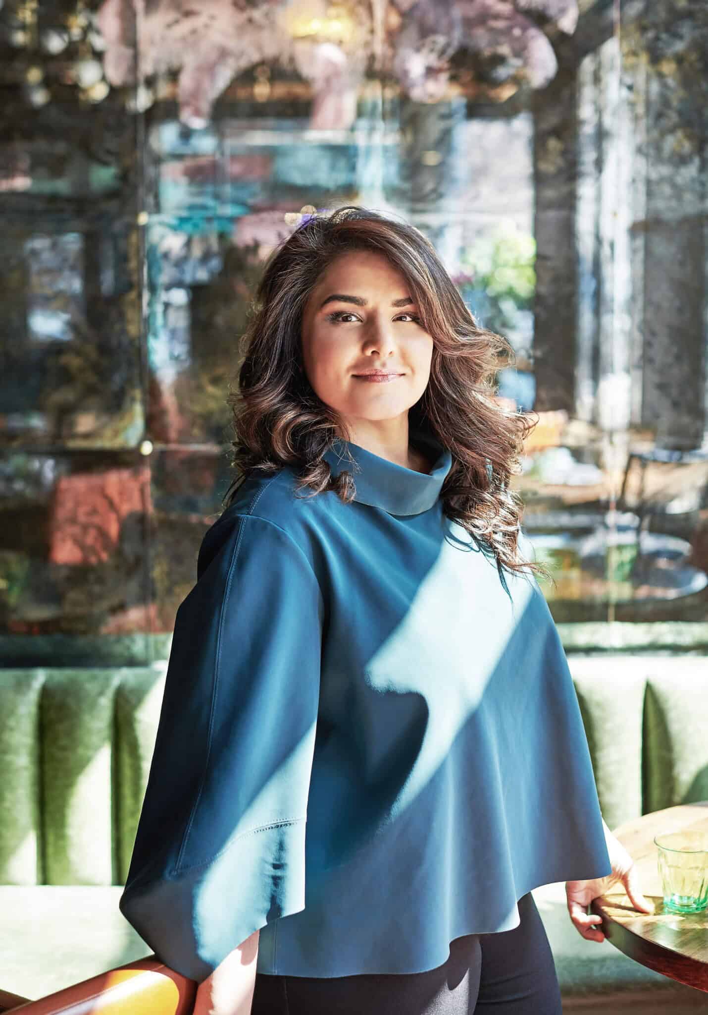 A woman with long wavy hair wearing a loose blue top stands indoors, smiling confidently at the camera. Sunlight filters through windows behind her, creating a warm and inviting atmosphere.