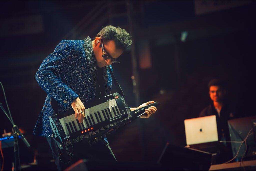 A musician wearing sunglasses and a blue patterned jacket plays a keytar on stage under colorful lights, while another person sits in the background near a laptop.