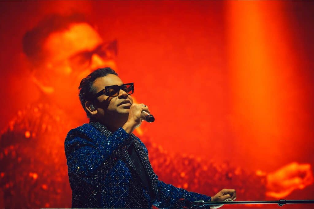 A man in a sparkly blue jacket and sunglasses sings into a microphone on stage, with his large, blurred image projected on a red-lit background behind him.