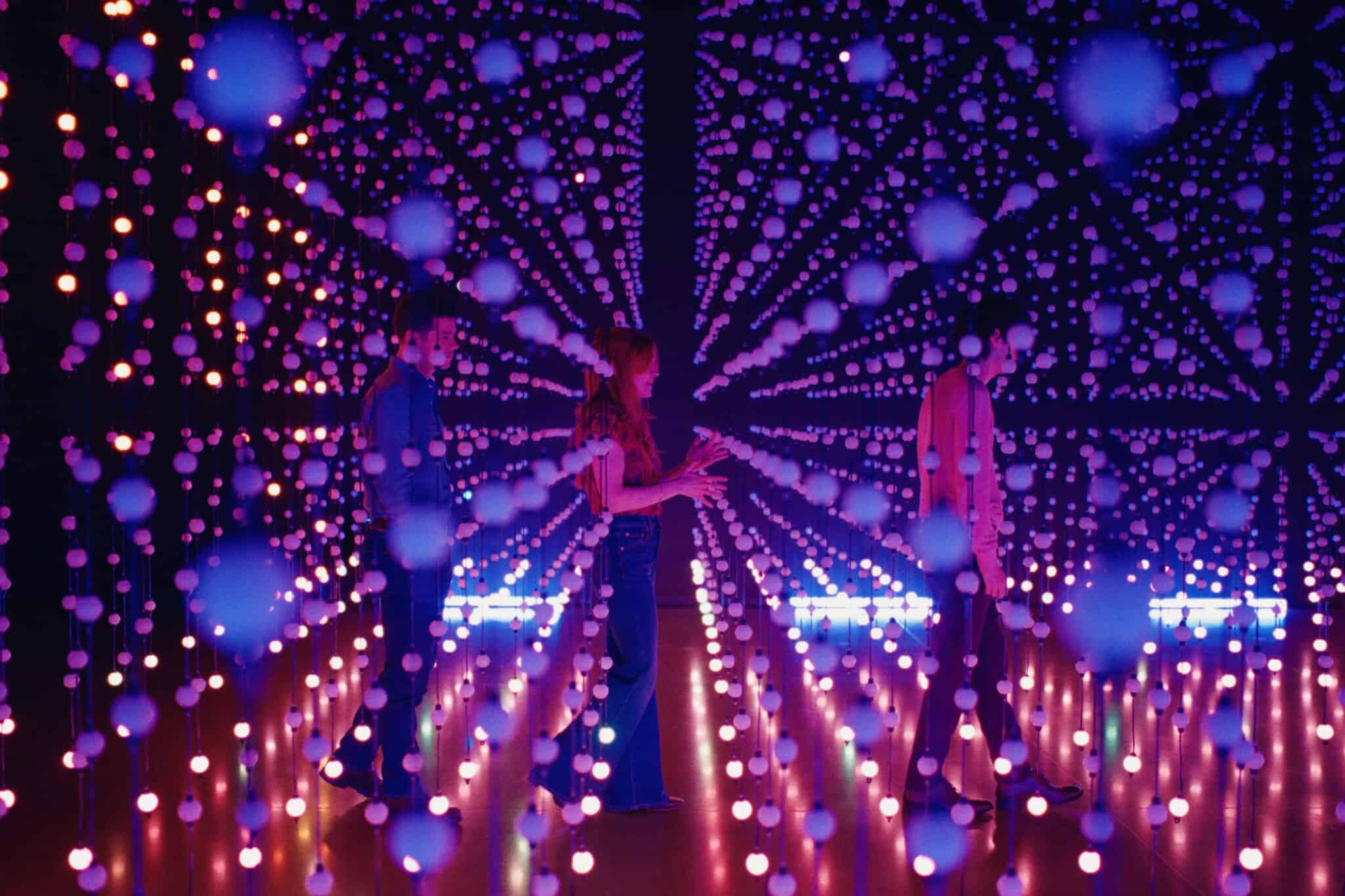 Three people stand inside a dark room filled with hanging strings of glowing purple and orange lights, creating a tunnel-like, immersive, and futuristic visual effect.