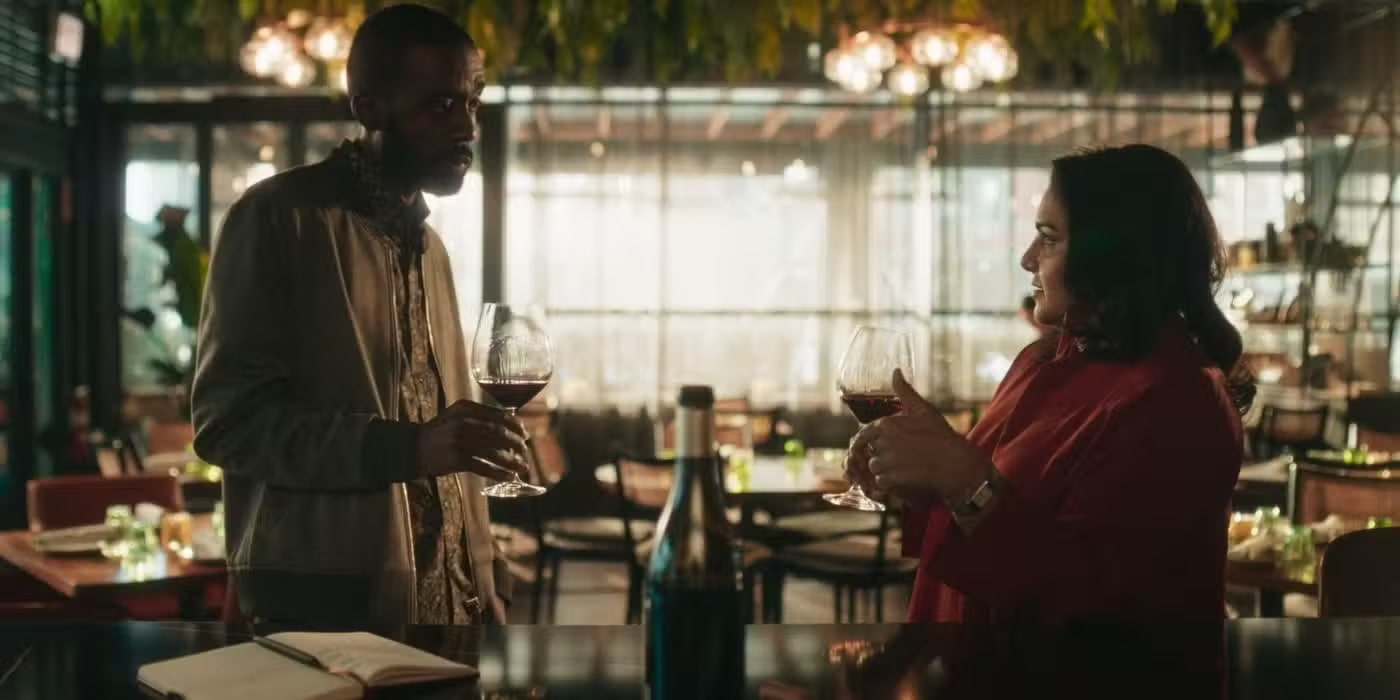 Two people stand at a restaurant bar, each holding a glass of red wine and talking. A wine bottle and open notebook are on the counter; tables and hanging lights are visible in the softly lit background.
