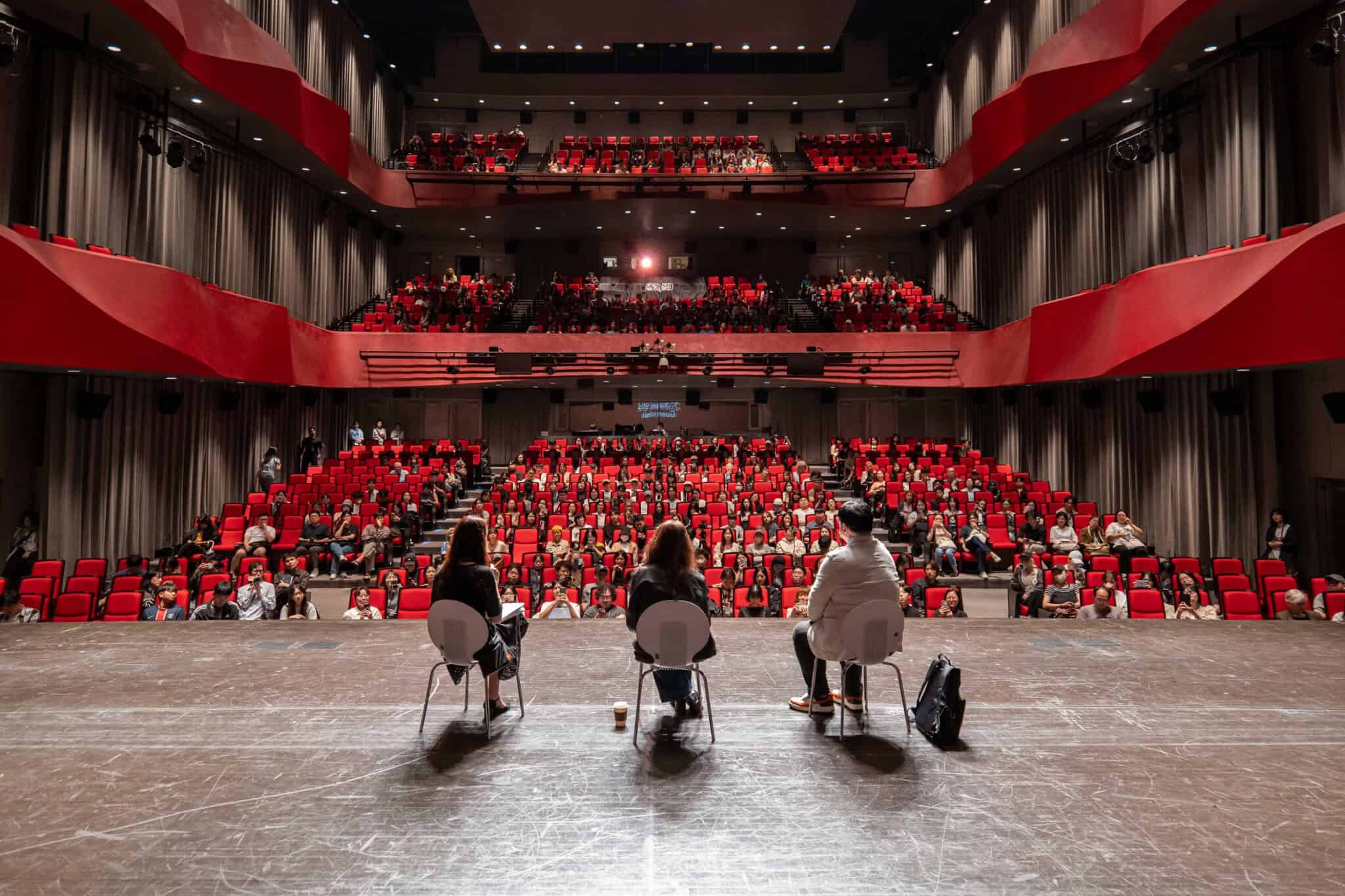 Three people sit on stage facing an audience in a modern auditorium filled with red seats. The audience is watching attentively. The viewpoint is from behind the speakers, looking out at the crowd.