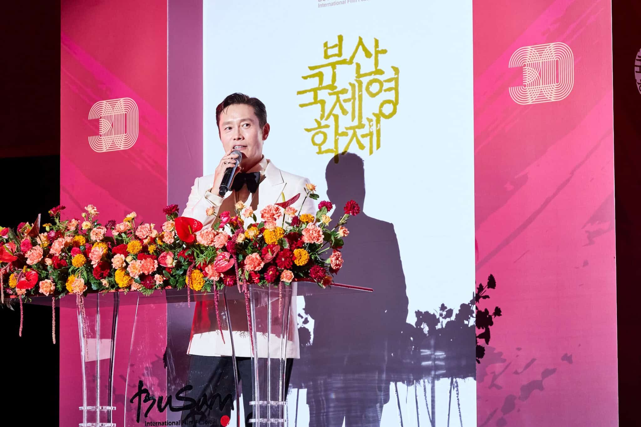 A man in a white suit speaks at a podium decorated with colorful flowers, with the Busan International Film Festival logo displayed on a screen behind him.