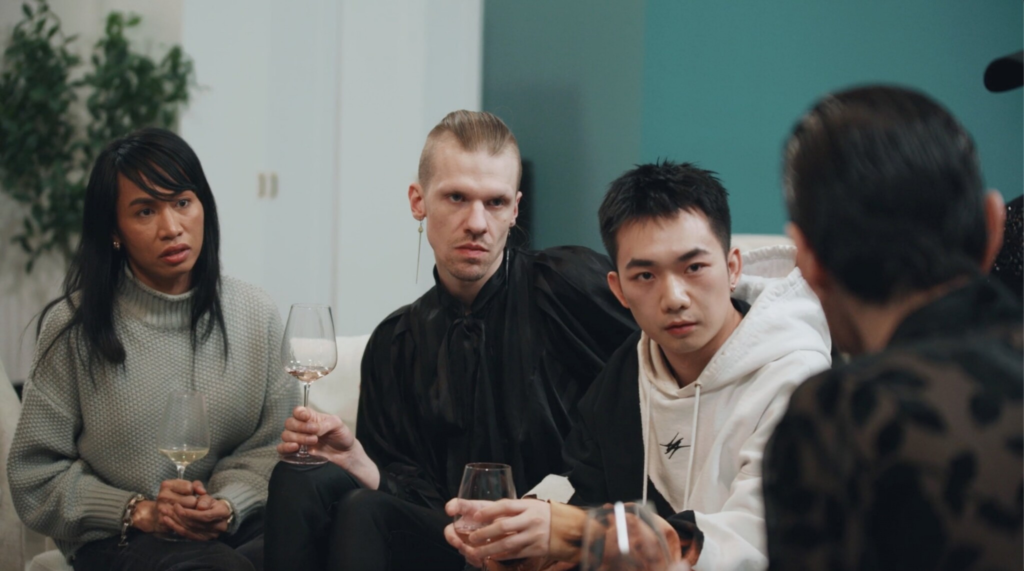 Four people sit closely together having a serious conversation. Three hold wine glasses and listen intently to the person opposite, with neutral or concerned expressions. There’s a plant and a teal wall in the background.