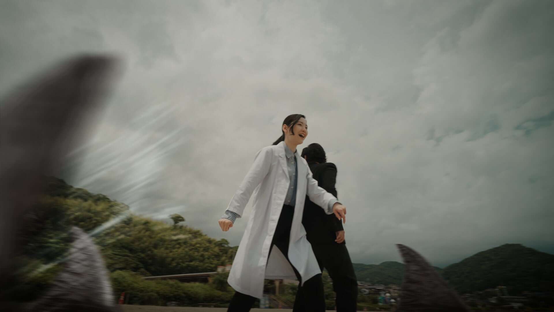 Two people, one in a white lab coat and the other in a dark suit, stand outdoors with dramatic clouds overhead. Large, blurred shark fins appear in the foreground, creating a sense of tension and danger.