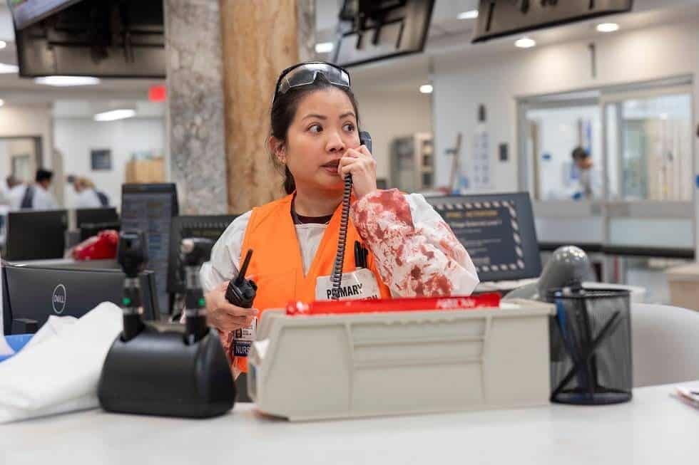 A woman in safety gear stands at a desk in a busy office or hospital, holding a walkie-talkie and speaking into a microphone, appearing alert and focused. Monitors and office supplies are visible on the desk.