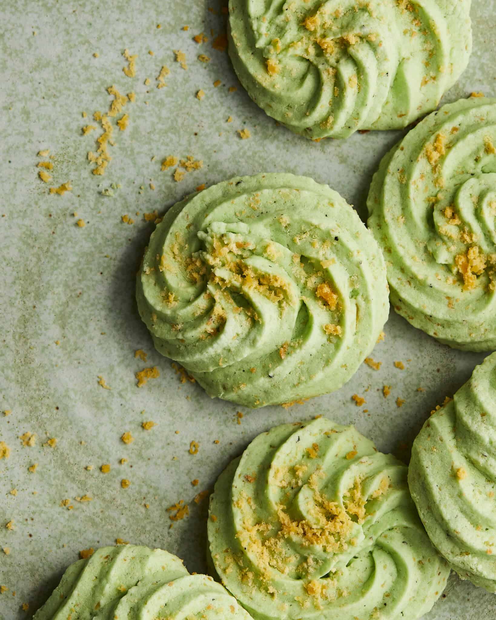 Green swirled cookies topped with crumbs are arranged on a parchment-lined surface, with a few crumbs scattered around.