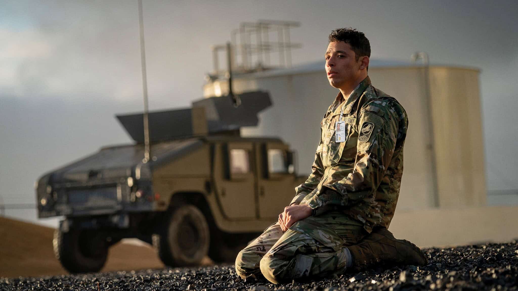 A person in military uniform kneels on rocky ground outdoors, with a military vehicle and large storage tank in the background under a cloudy sky.