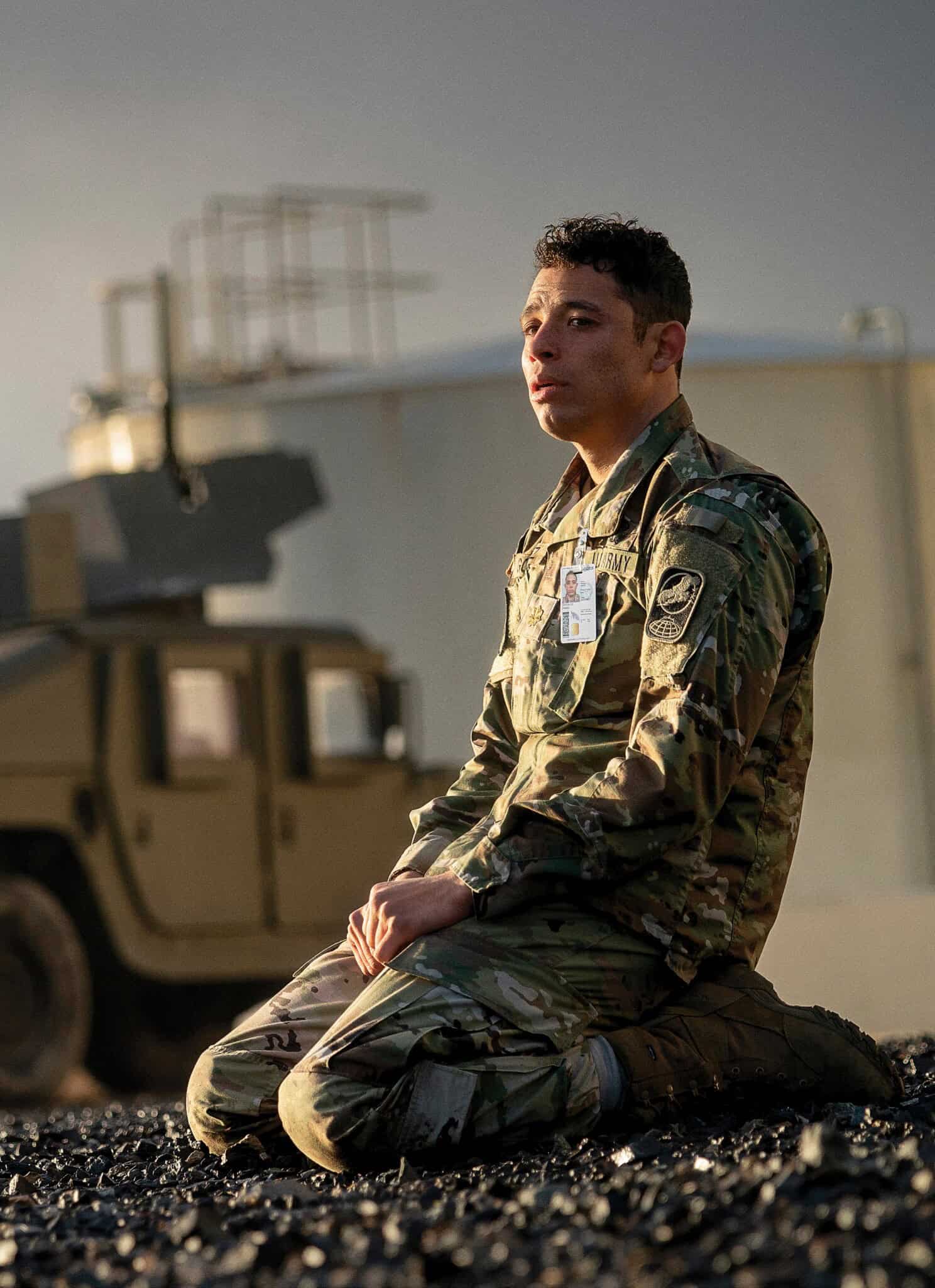 A person in military uniform kneels on rocky ground outdoors, with a military vehicle and large storage tank in the background under a cloudy sky.