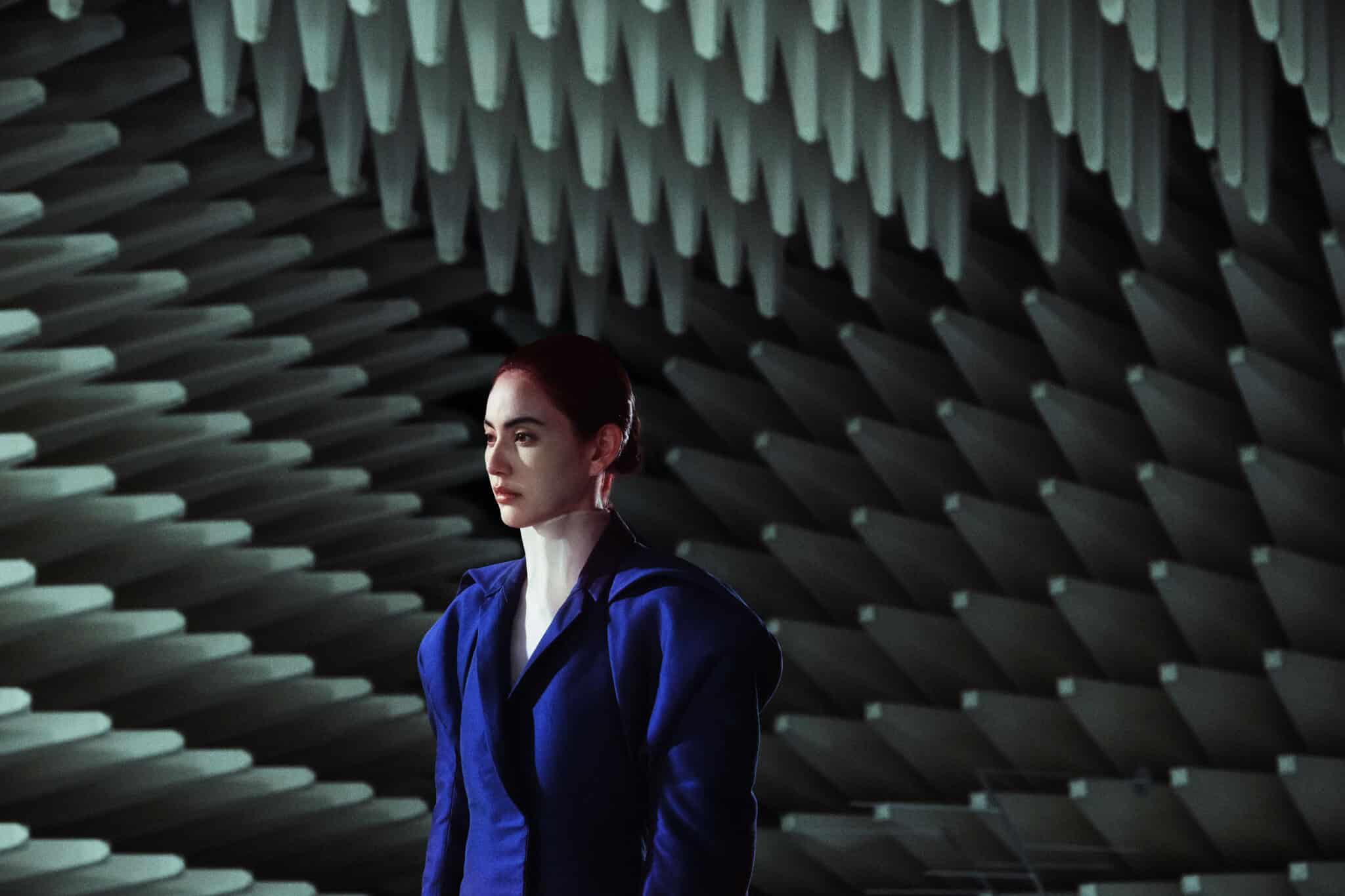 A person wearing a futuristic blue outfit stands in front of a wall covered with large, pointed acoustic foam panels, creating a geometric and modern atmosphere.