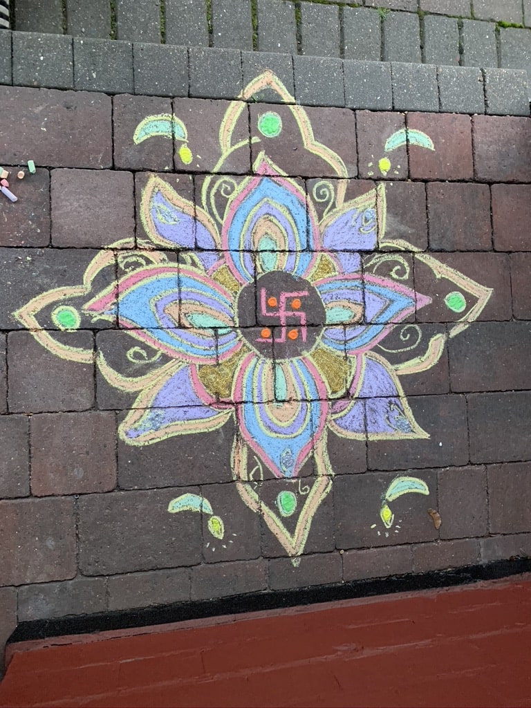 Colorful rangoli design drawn with chalk on brick pavement, featuring symmetrical flower petals in purple, blue, green, and yellow, with a red swastika symbol in the center and decorative accents around it.