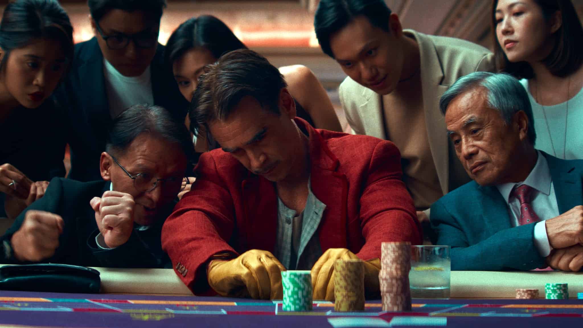A tense group of people gather around a casino table, focused on a man in a red jacket and yellow gloves handling poker chips, while others watch anxiously, anticipating the outcome.