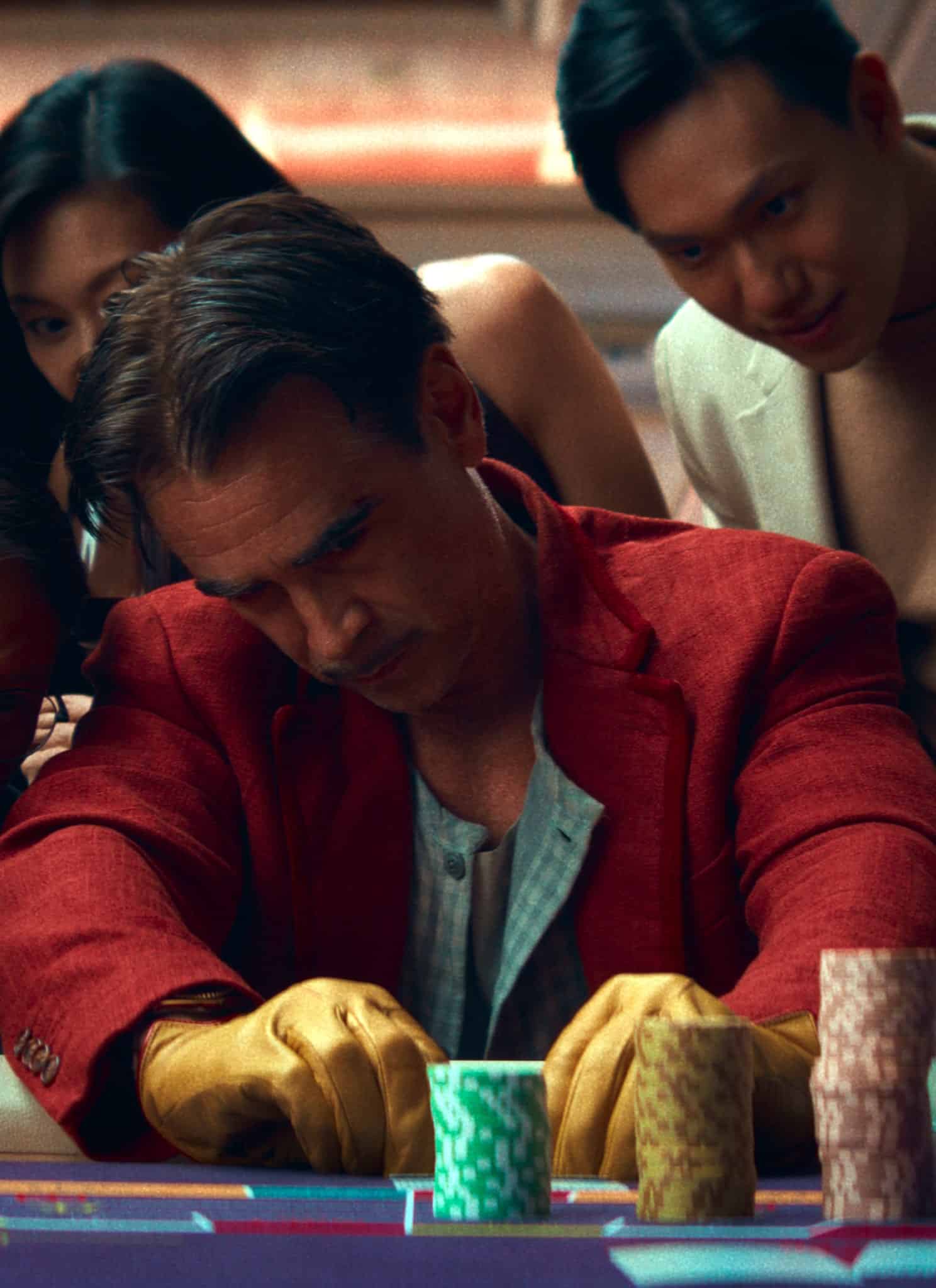 A tense group of people gather around a casino table, focused on a man in a red jacket and yellow gloves handling poker chips, while others watch anxiously, anticipating the outcome.