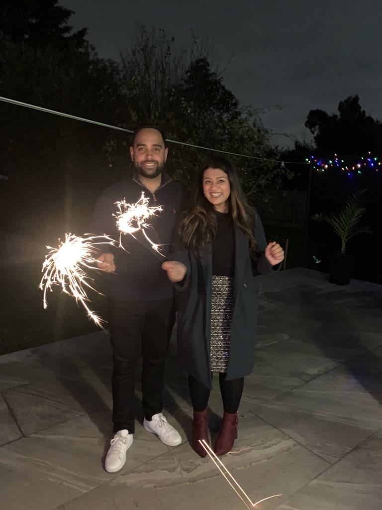 Two people stand outside at night, smiling and holding sparklers. They are on a patio with colorful string lights in the background and trees surrounding the area.