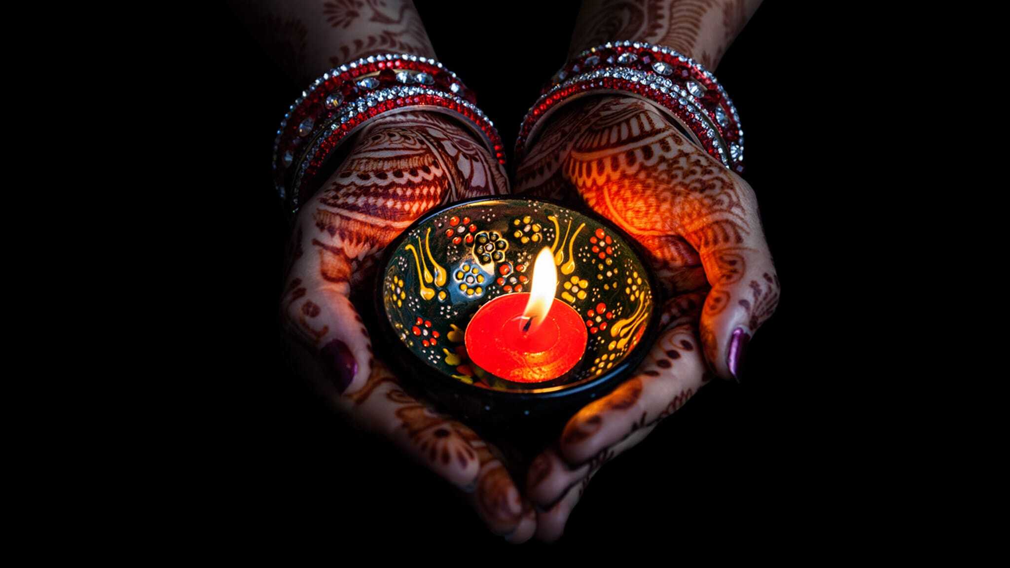 Hands adorned with henna and red bangles holding a decorated clay lamp with a burning red candle, glowing warmly against a dark background.