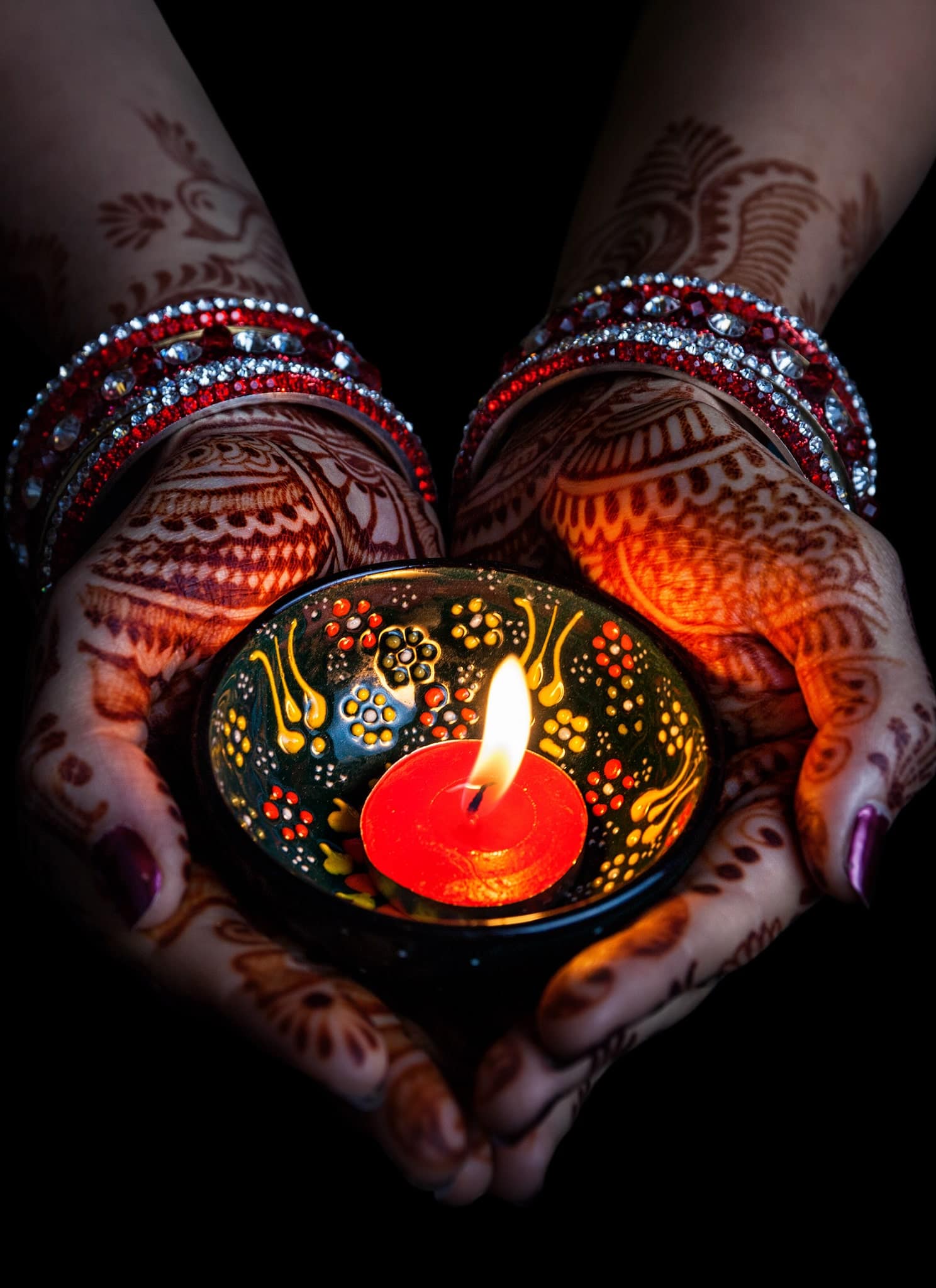 Hands adorned with henna and red bangles holding a decorated clay lamp with a burning red candle, glowing warmly against a dark background.