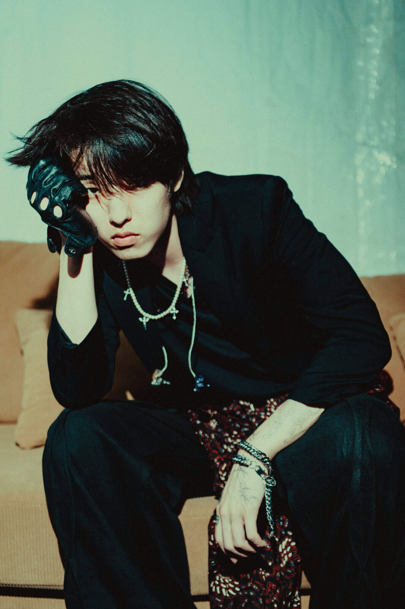 A person with medium-length dark hair sits on a sofa, wearing a black outfit, patterned pants, and a fingerless glove. Their hand rests on their face, and they have a contemplative expression.