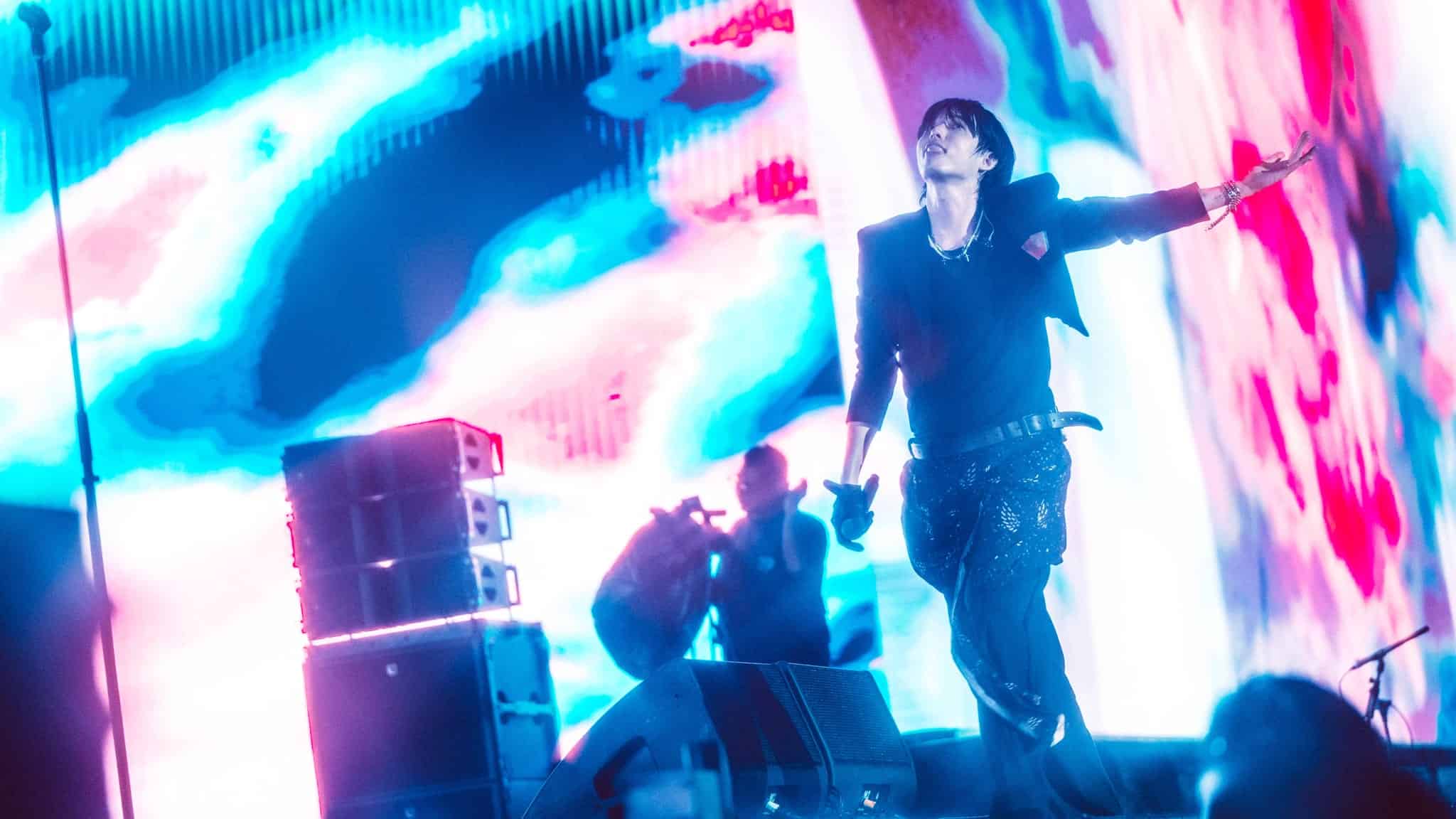 A performer in dark clothing energetically sings on stage with one arm outstretched, set against a colorful, abstract background of blue, pink, and white lights. Stage equipment and a photographer are visible in the background.