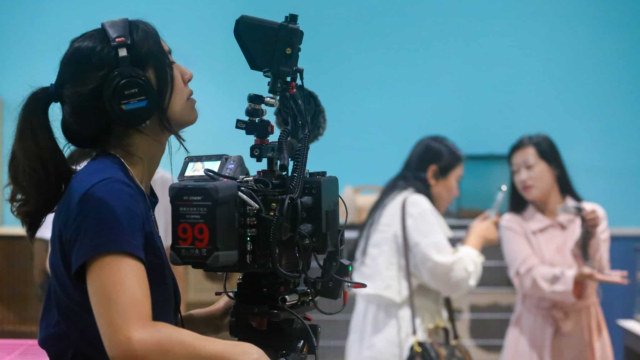 A woman wearing headphones operates a professional video camera, focusing on two women in the background who are engaged in conversation, with one gesturing and the other holding a phone, against a bright blue wall.
