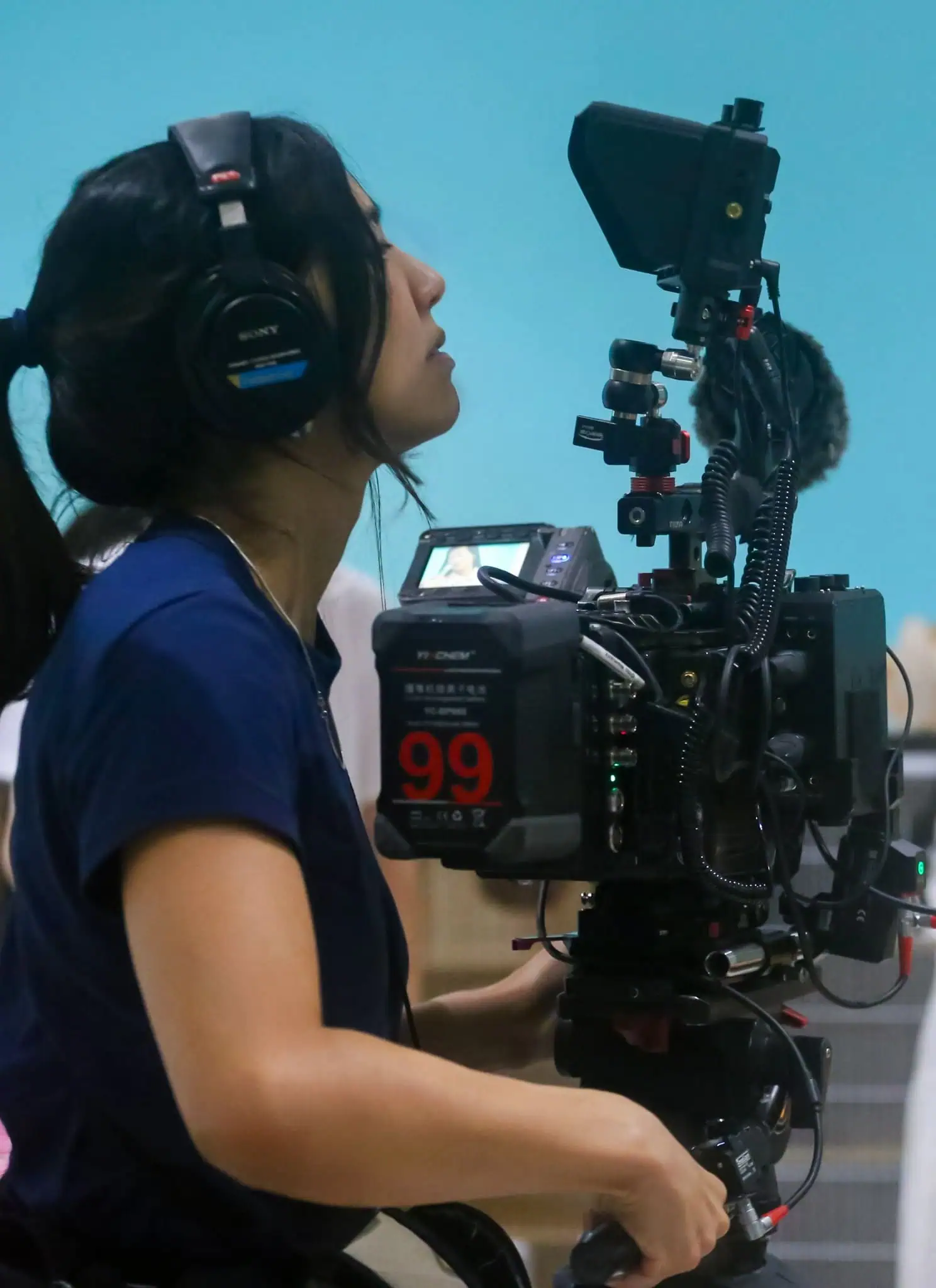 A woman wearing headphones operates a professional video camera, focusing on two women in the background who are engaged in conversation, with one gesturing and the other holding a phone, against a bright blue wall.