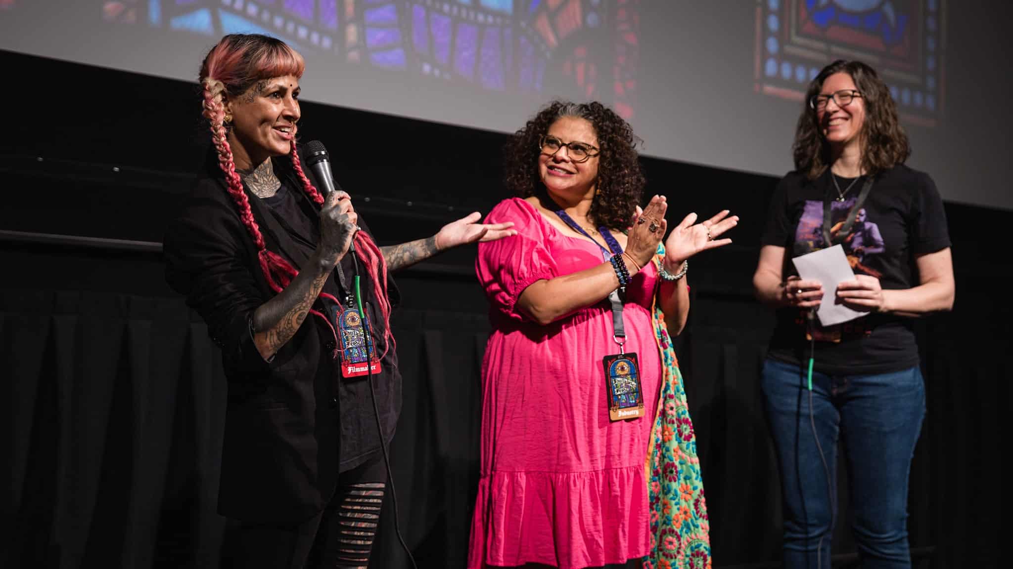 Three women stand on a stage in front of a projected stained glass image; one with pink braids speaks into a microphone, the second in a pink dress claps, and the third smiles while holding a piece of paper.