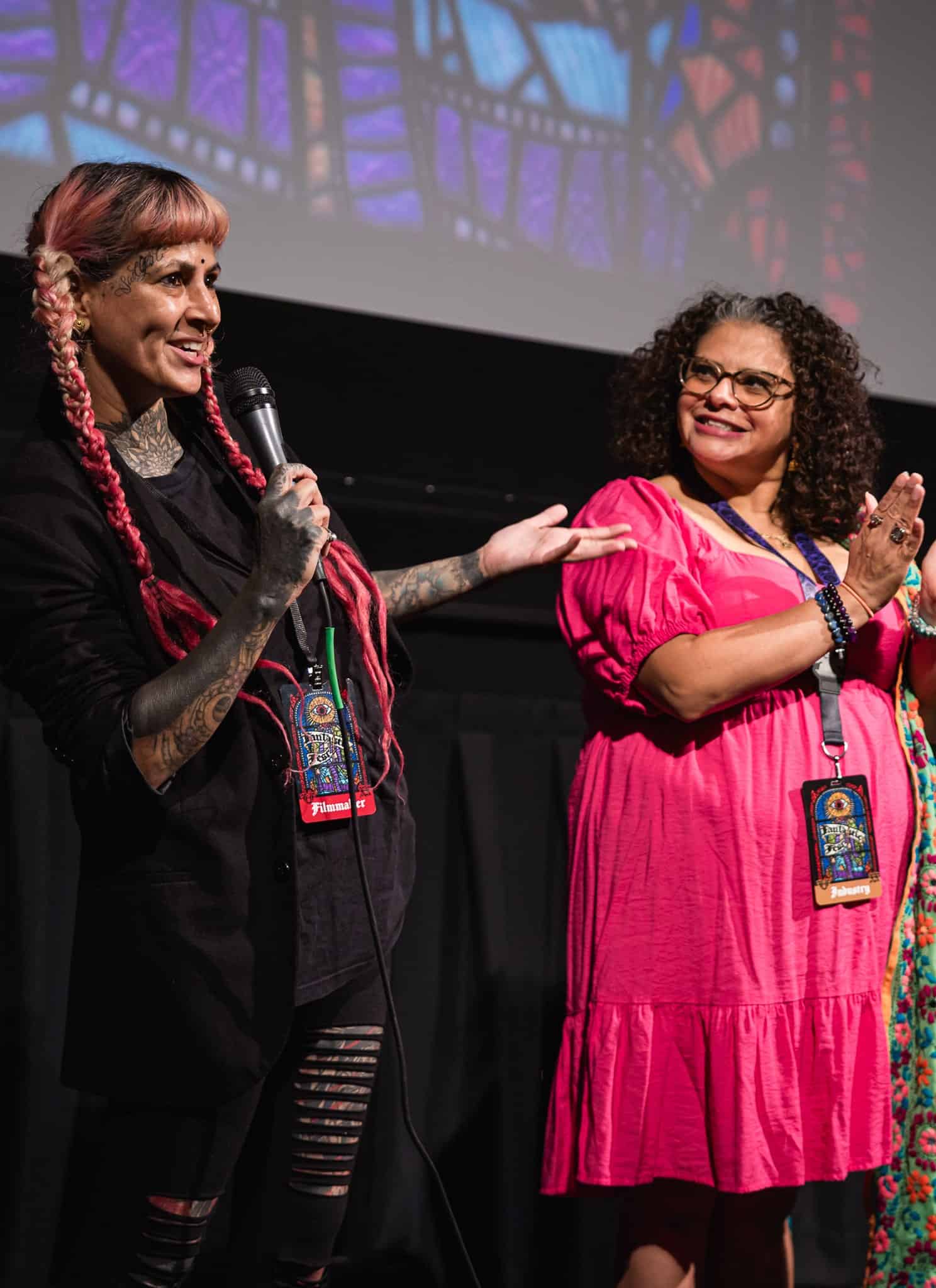 Three women stand on a stage in front of a projected stained glass image; one with pink braids speaks into a microphone, the second in a pink dress claps, and the third smiles while holding a piece of paper.
