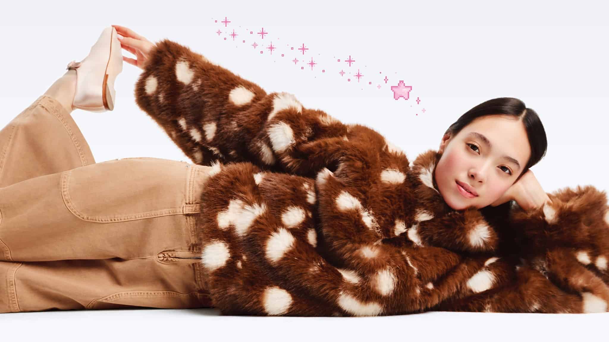A woman in a brown and white polka dot faux fur coat and tan pants lies on her side, propping up her head with one hand and holding her foot with the other, with pink sparkles and a star illustrated behind her.
