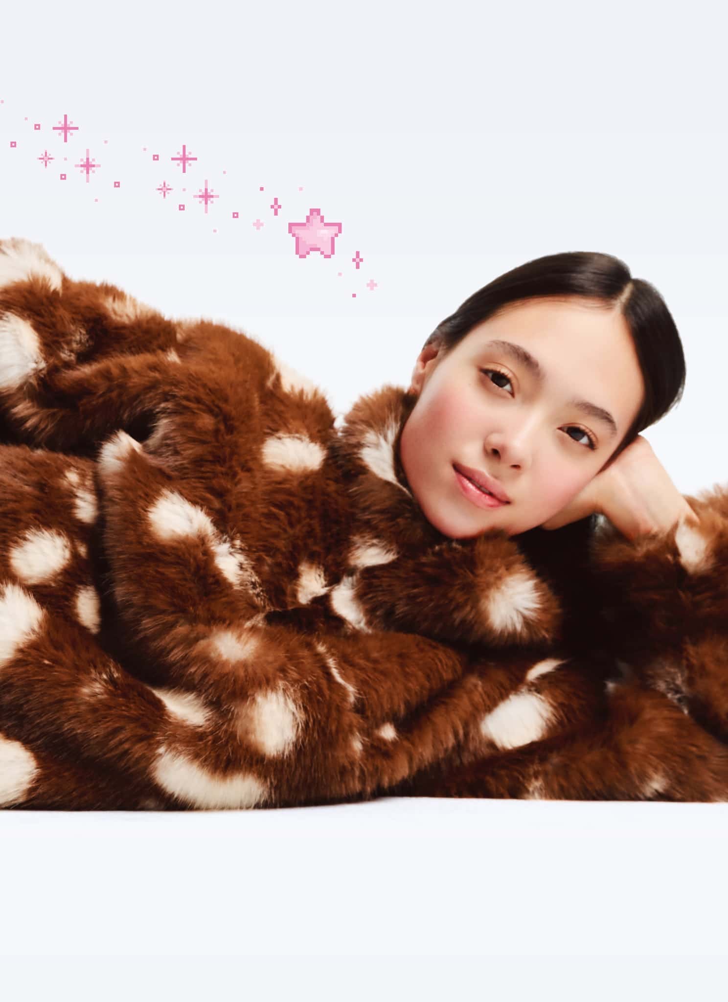 A woman in a brown and white polka dot faux fur coat and tan pants lies on her side, propping up her head with one hand and holding her foot with the other, with pink sparkles and a star illustrated behind her.
