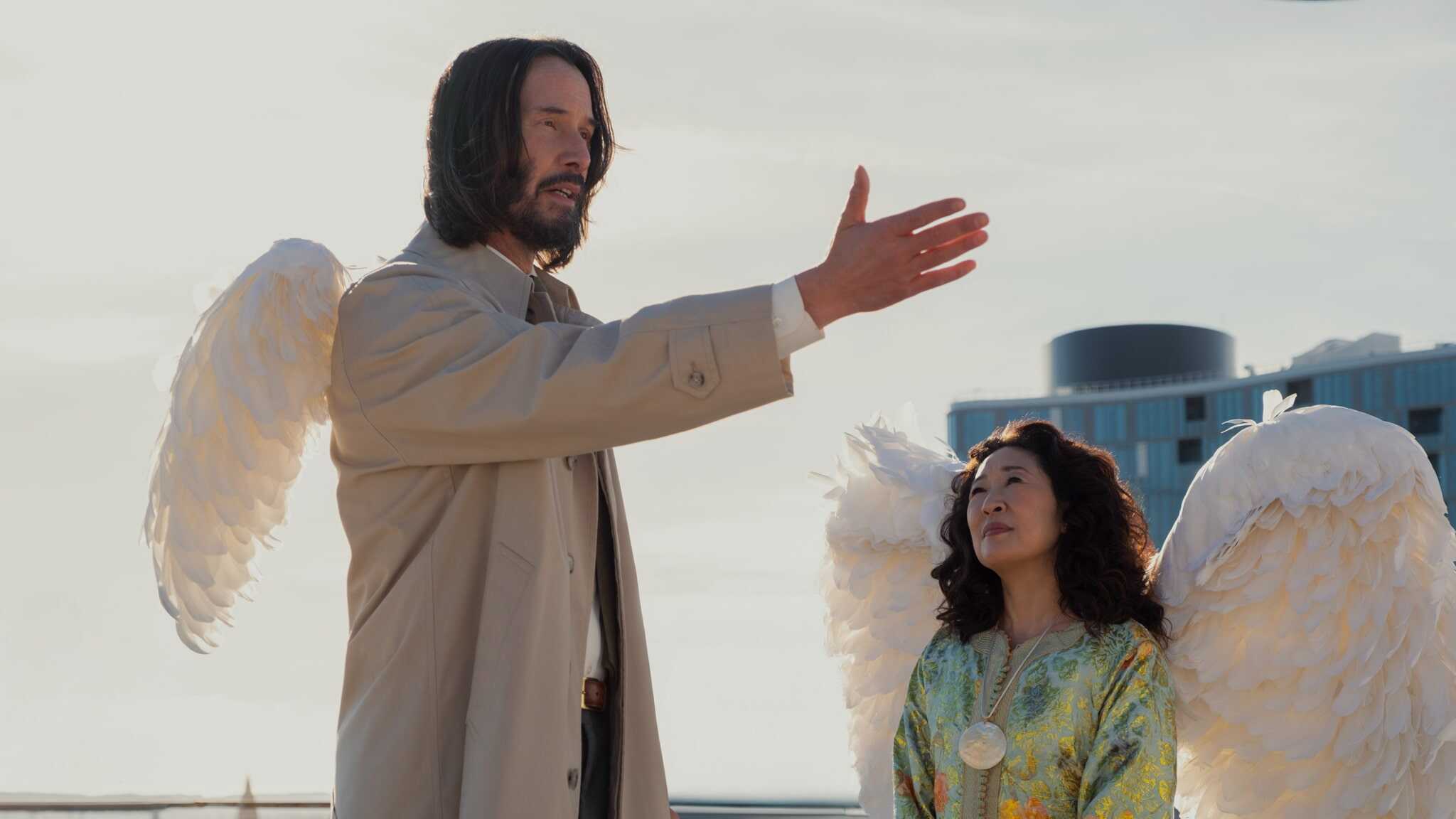 Two people dressed as angels with large white wings stand outdoors. One, with long hair and a beige coat, gestures with his hand while the other, with curly hair and a patterned outfit, looks up at him. A modern building is in the background.