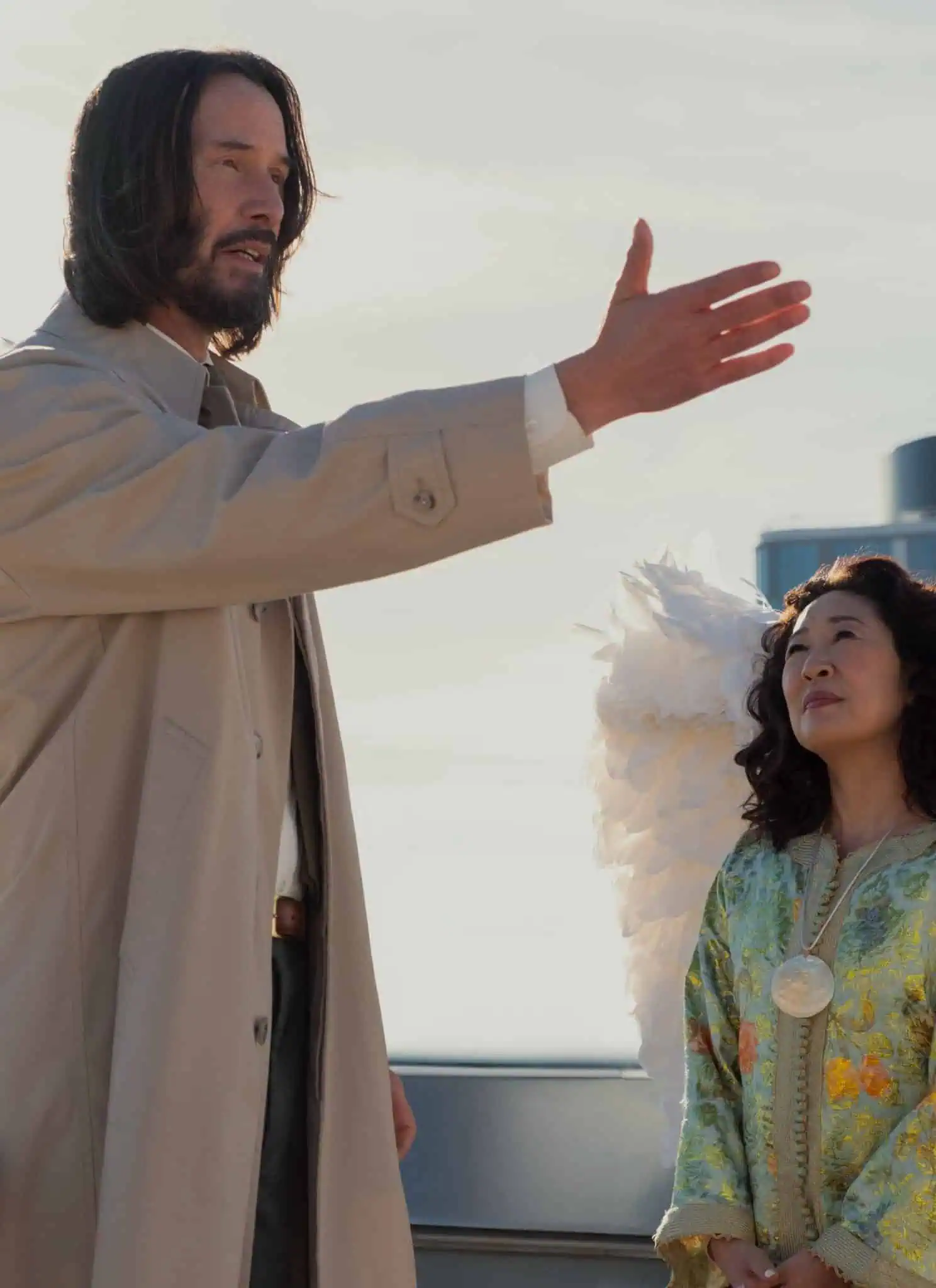 Two people dressed as angels with large white wings stand outdoors. One, with long hair and a beige coat, gestures with his hand while the other, with curly hair and a patterned outfit, looks up at him. A modern building is in the background.