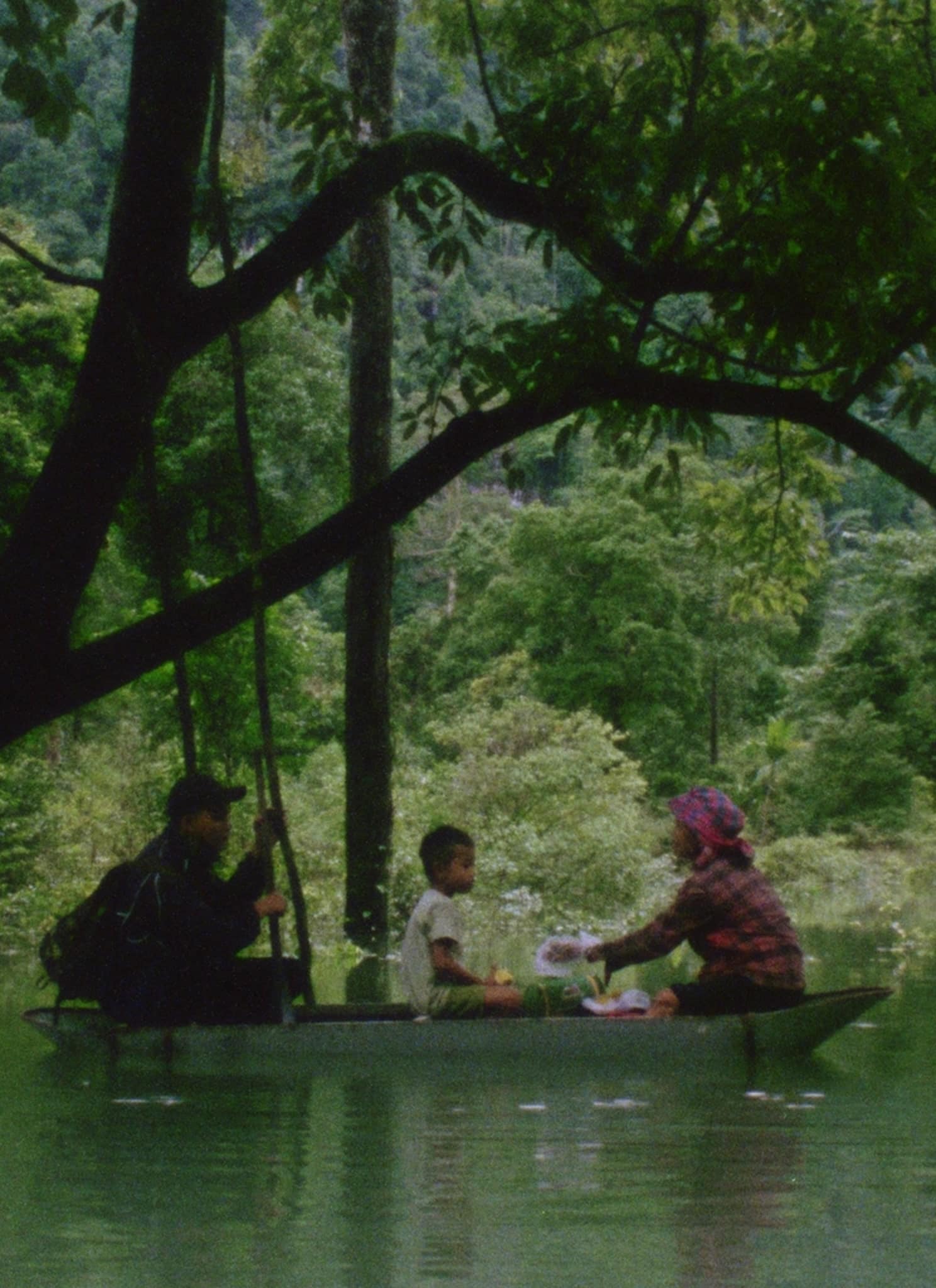 Three people sit in a small boat on a calm, green river surrounded by lush trees. The scene is peaceful, with one child and two adults, one paddling and the other interacting with the child.