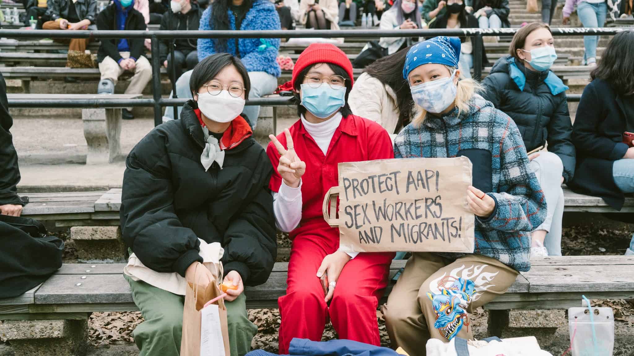 Three people sit on benches outdoors, all wearing face masks. The person in the middle holds a sign that reads Protect AAPI sex workers and migrants! Others sit in the background, also wearing masks.