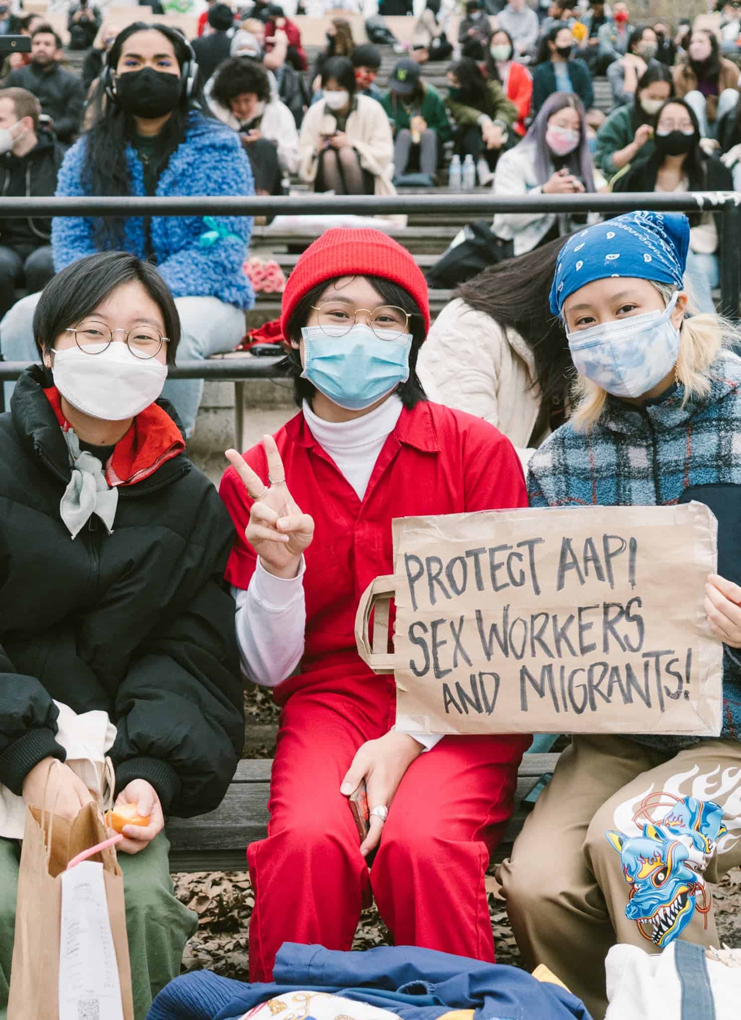 Three people sit on benches outdoors, all wearing face masks. The person in the middle holds a sign that reads Protect AAPI sex workers and migrants! Others sit in the background, also wearing masks.